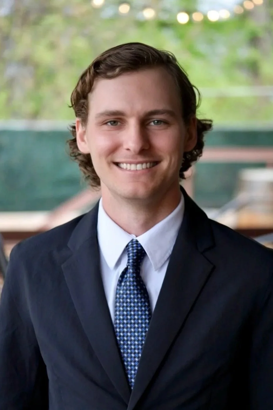 A young man with brown hair, wearing a dark suit, white shirt, and patterned tie, smiling at the camera with a blurred background of trees and outdoor scenery.