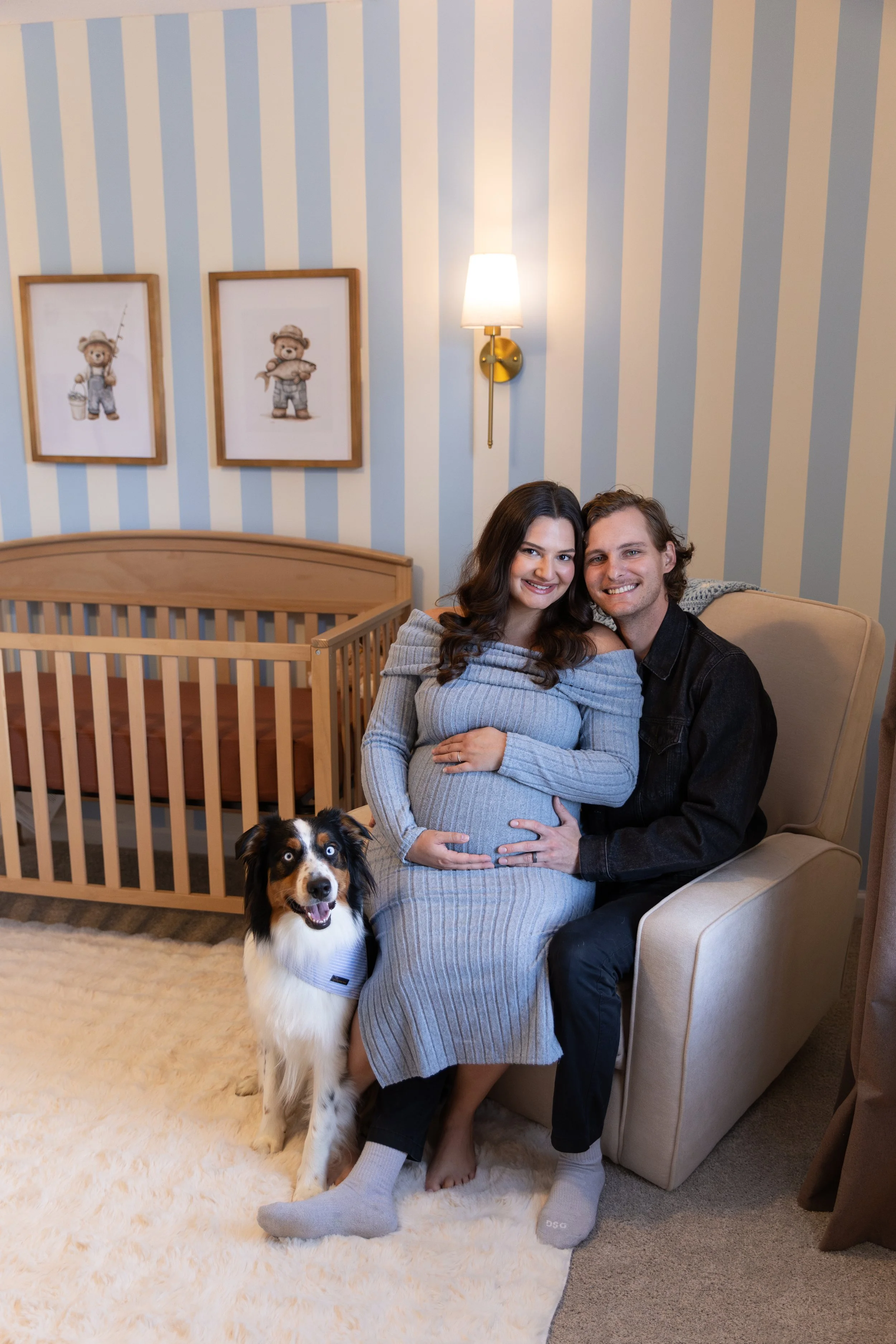 A pregnant woman, a man, and a black and white dog in a cozy nursery with blue and white striped wallpaper, framed bear artwork, and a wooden crib.