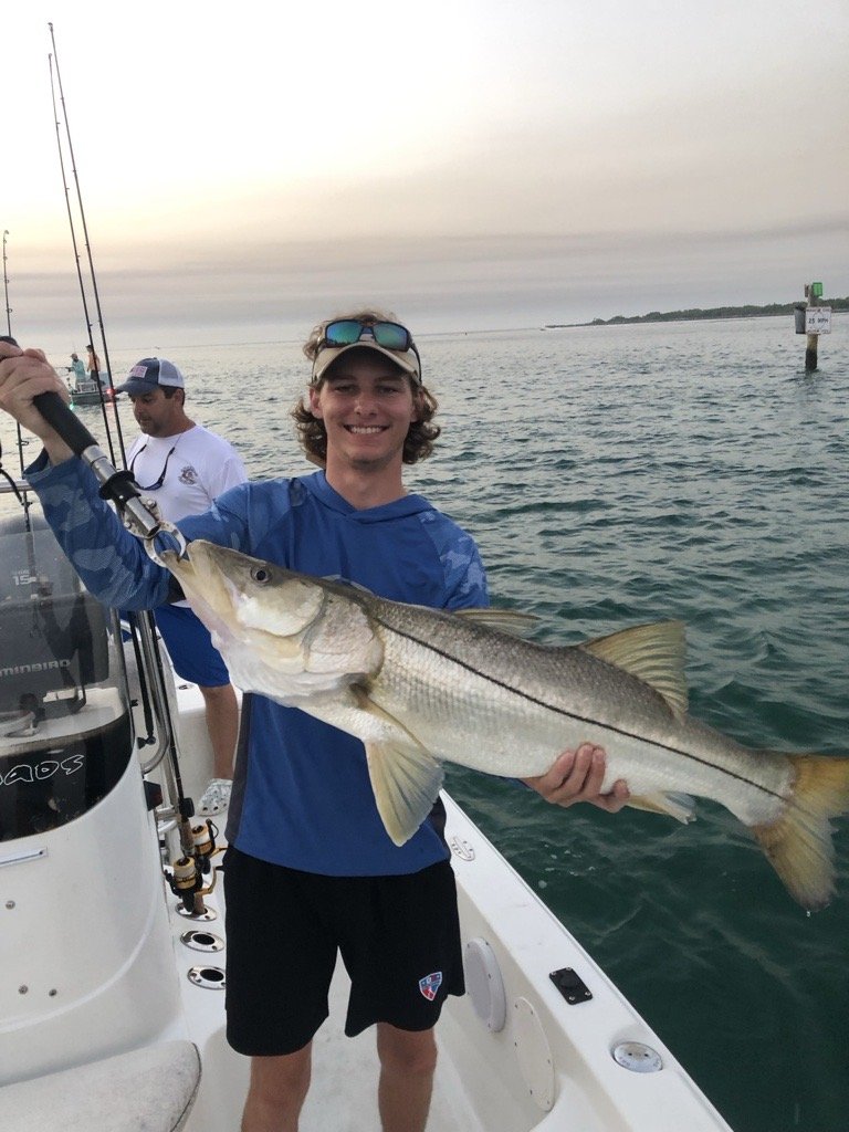 Young man in a blue jacket and black shorts holding a large fish on a boat, smiling, with another person in the background, calm water, and a sunset sky.