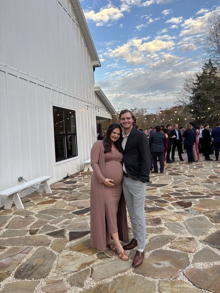 A pregnant woman and a man smiling at the camera during an outdoor event with string lights and other people in the background.