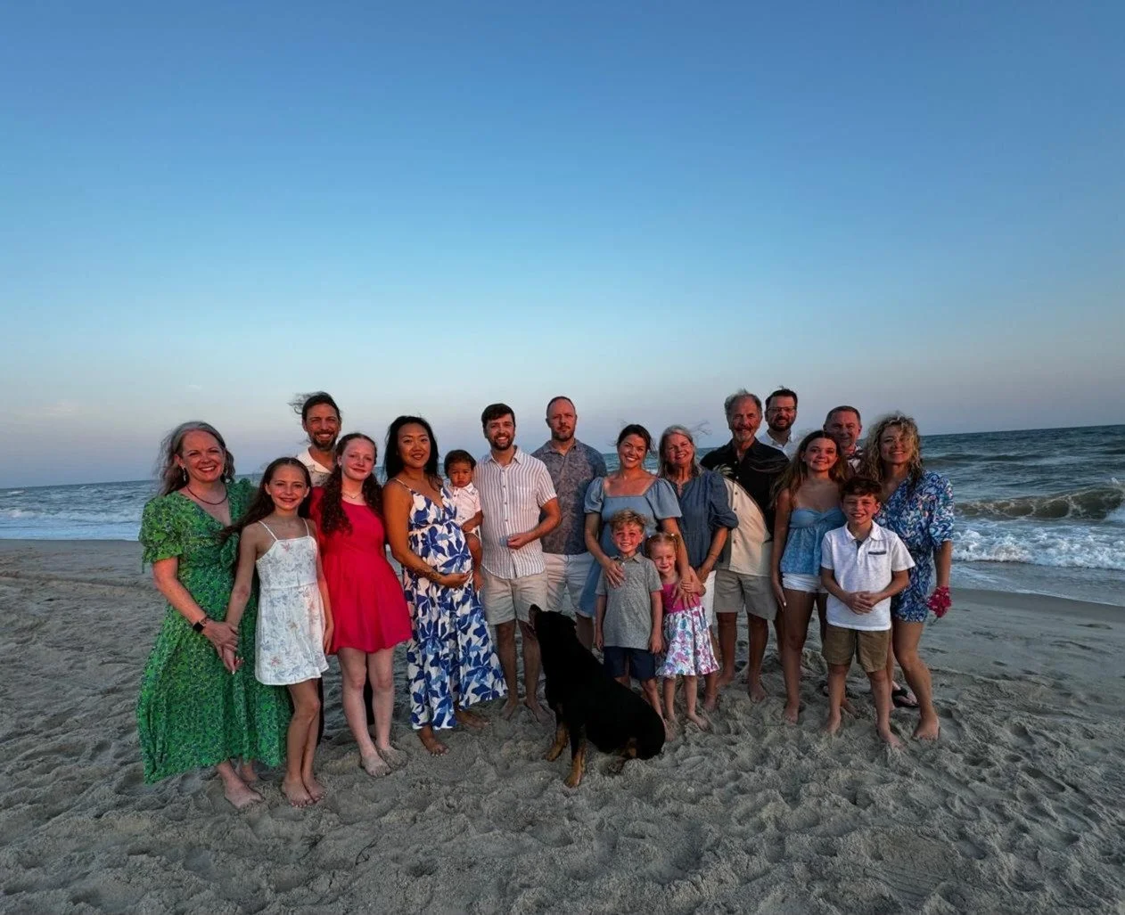 A large family group stands on the beach with the ocean and sky in the background, during sunset, smiling at the camera.