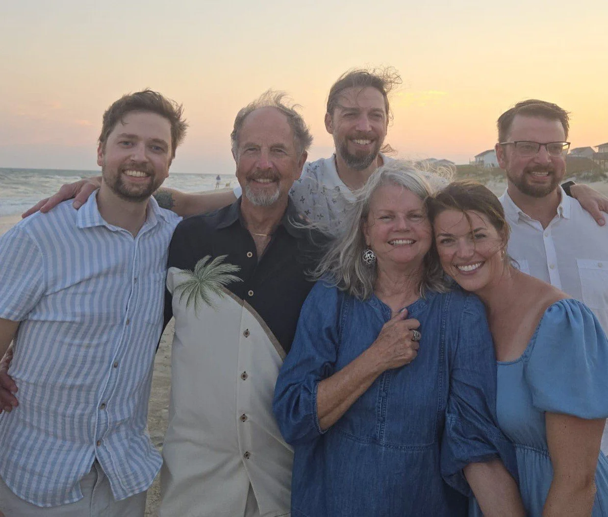 Group of six people smiling on the beach during sunset, with ocean waves and houses in the background.