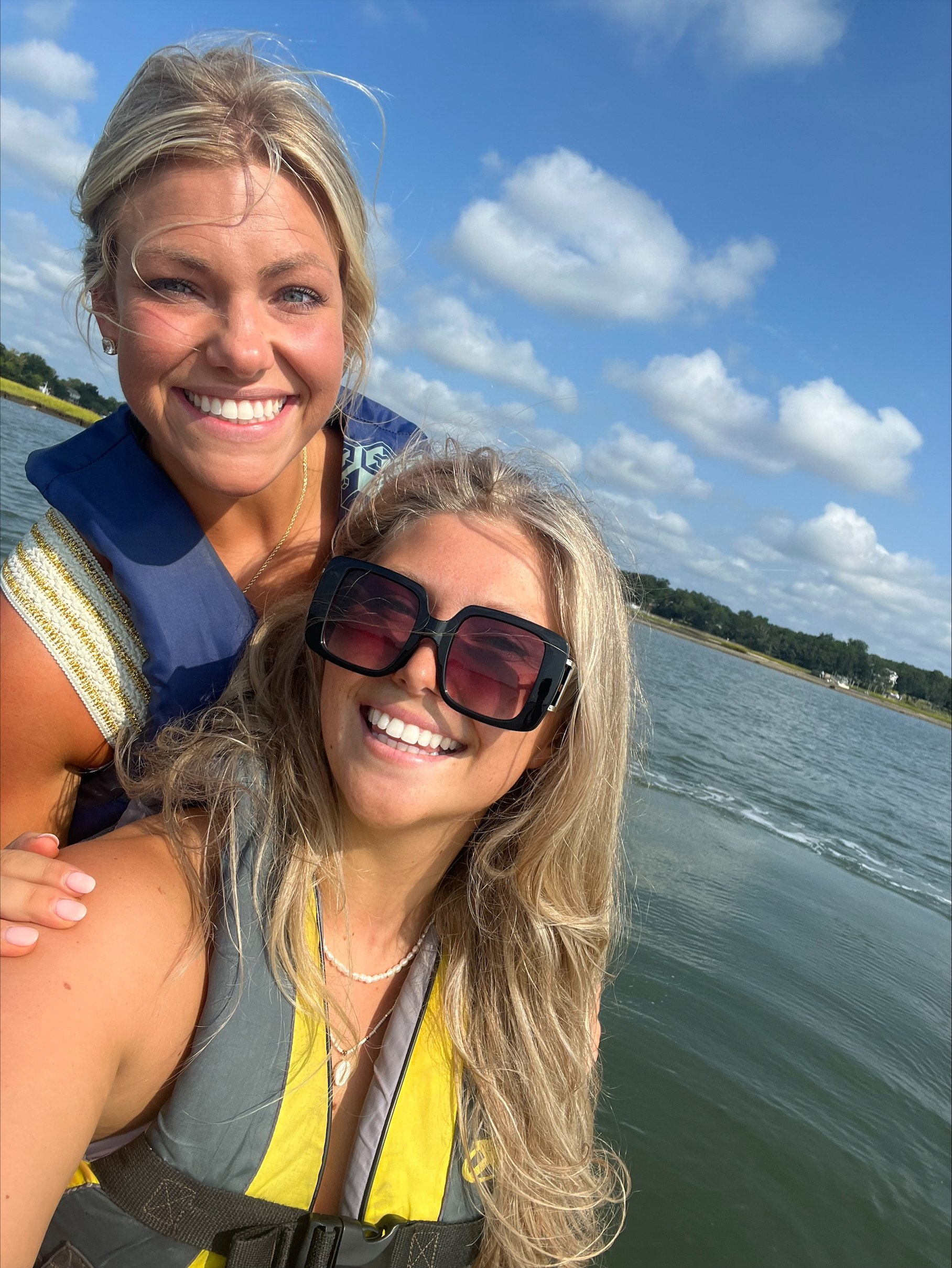 Two women smiling for a selfie on a boat or watercraft, one wearing large sunglasses and a life jacket, the other with blonde hair and a blue life jacket, with water and a partly cloudy sky in the background.