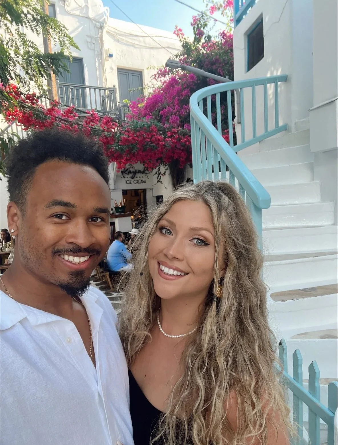 A smiling man and woman taking a selfie outdoors in front of white stairs and colorful pink and red bougainvillea flowers.