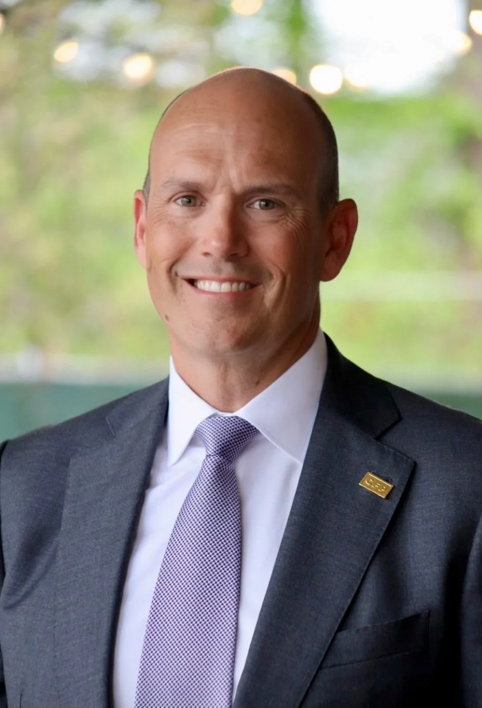 A smiling man in a business suit and tie, with a blurred green outdoor background.