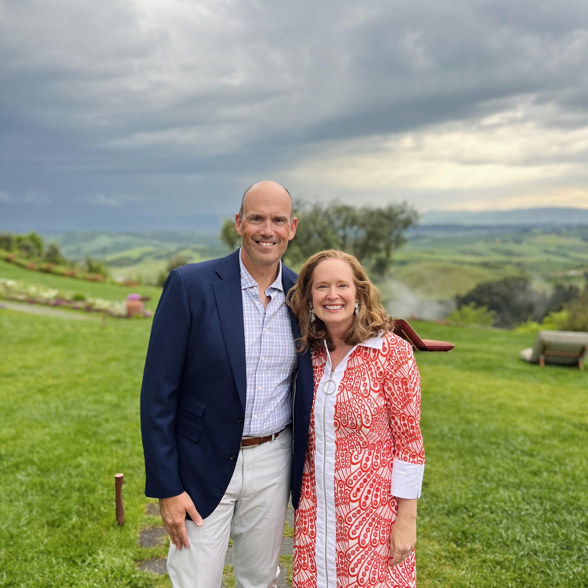 A man and woman standing outdoors in a scenic green landscape with rolling hills and trees, overcast sky, smiling at the camera.
