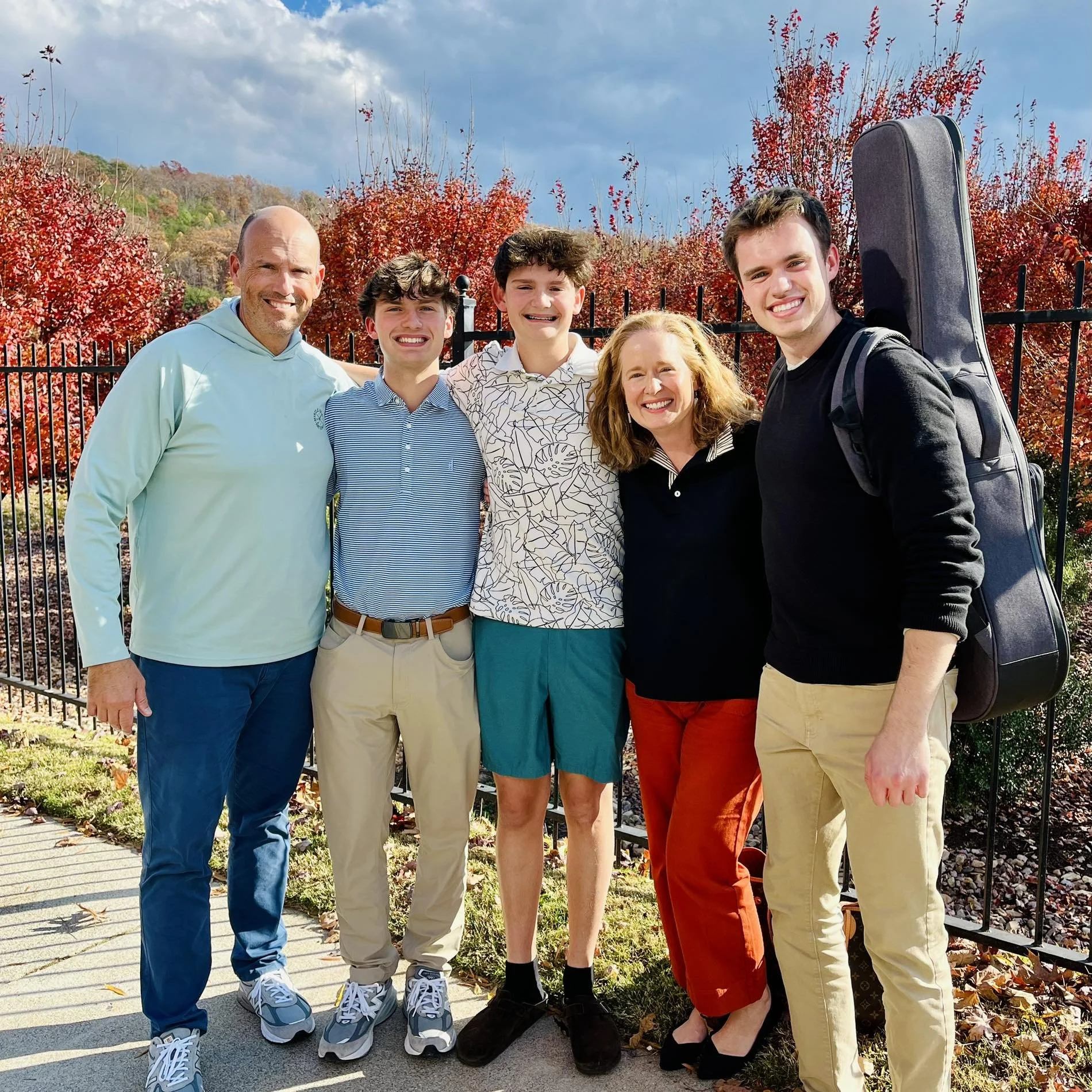 A family of six standing outside in front of red autumn trees, smiling at the camera.