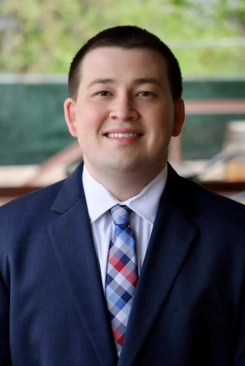 A young man dressed in a navy suit, white shirt, and a multicolored plaid tie, smiling in front of a blurred background with natural greenery.