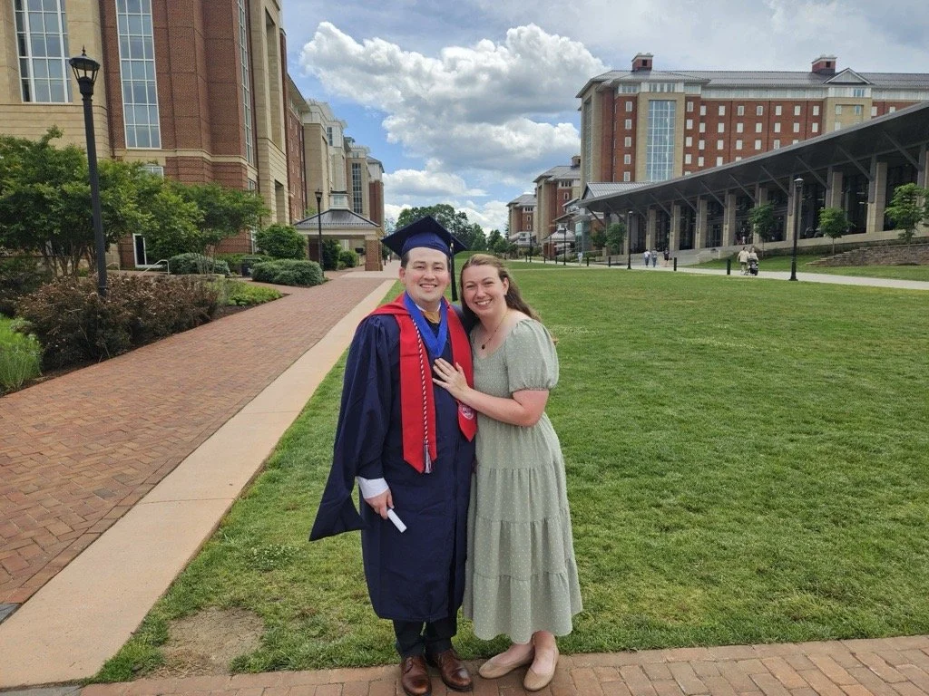 A young man in a graduation gown and cap standing next to a woman, both smiling, with a campus background featuring brick buildings, a walkway, picnic tables, and a partly cloudy sky.