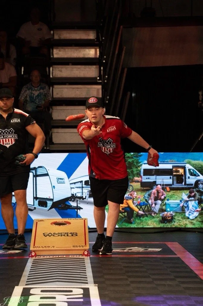 A young man in a red 'USA Forces' jersey and cap throwing a cornhole bean bag toward a wooden cornhole board during an indoor event. There are spectators in the background, and a large screen displaying an outdoor camping scene with a camper, a van, and people sitting around a campfire.