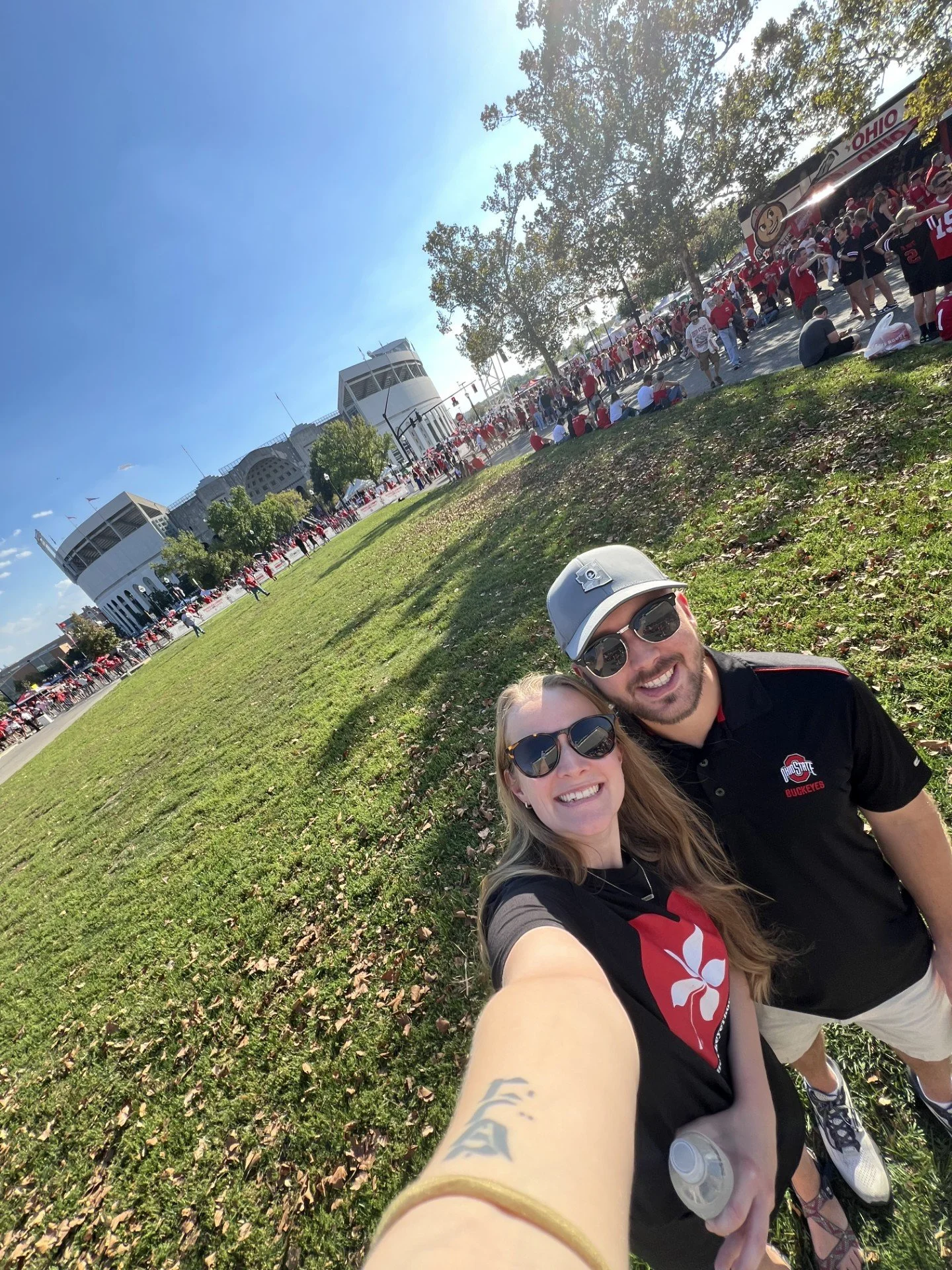 A smiling couple taking a selfie outdoors on a sunny day with a large crowd of people dressed in Ohio State Buckeyes apparel in the background. The woman has blonde hair, sunglasses, and a black shirt with a red and white logo. The man has a beard, sunglasses, a gray cap, and a black polo shirt with a red Ohio State logo.