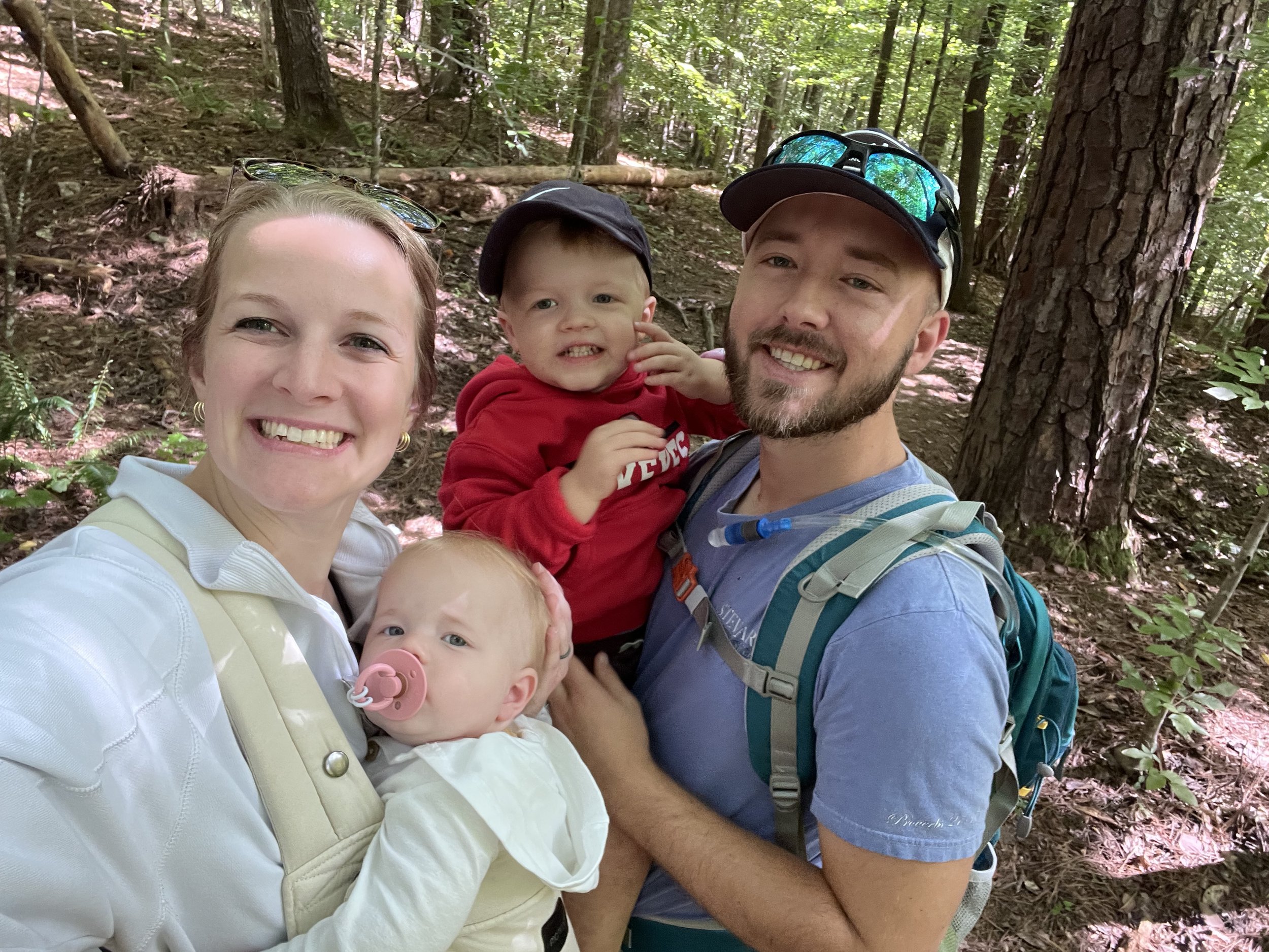 A family of four taking a selfie in a wooded forest during daytime. The mother has short blonde hair, and is smiling while holding a baby with a pacifier. The father, wearing a backpack and sunglasses on his head, is smiling and holding a toddler in a red hoodie. The forest has tall trees and green leaves, with dappled sunlight on the ground.