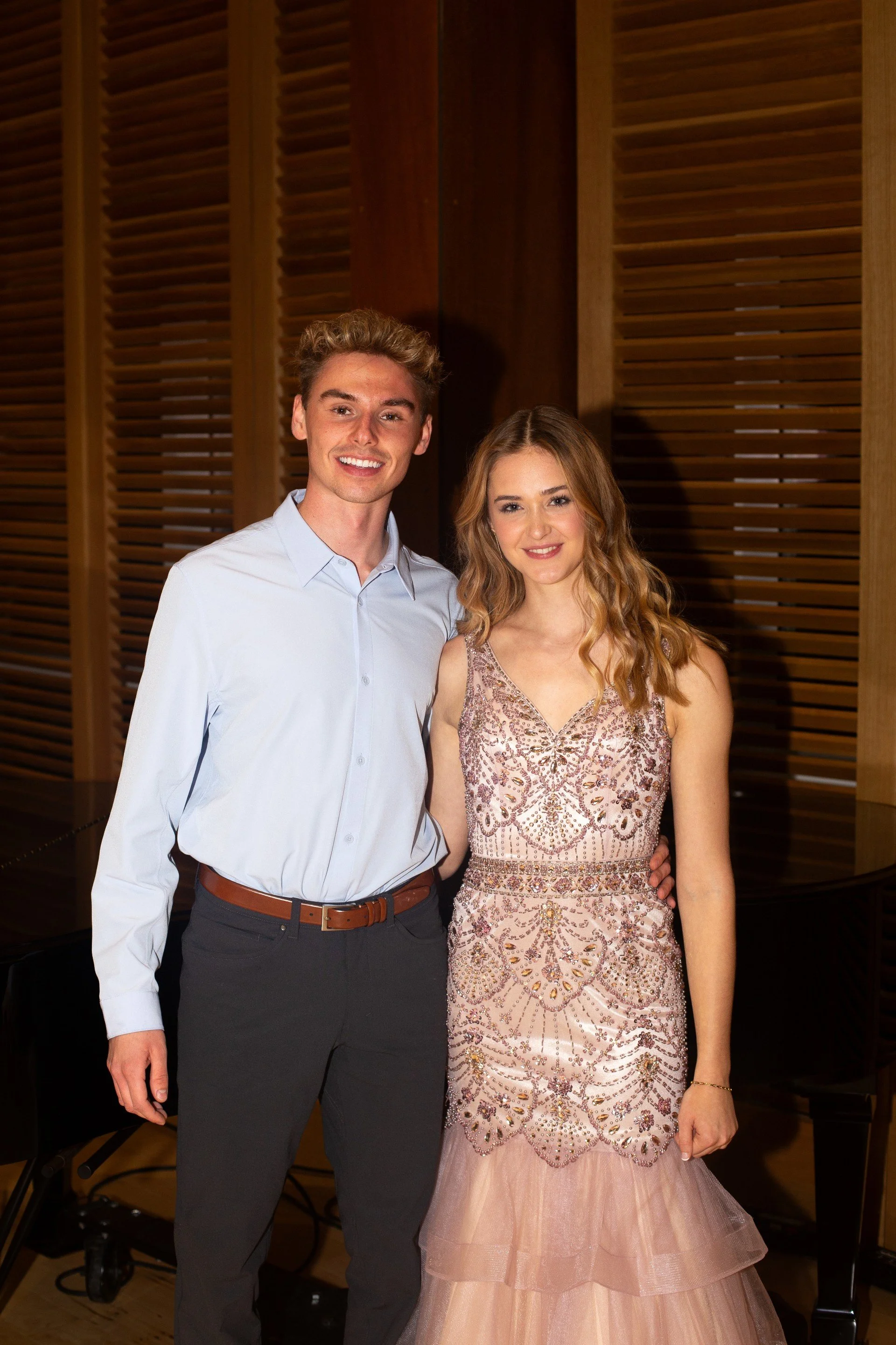 A young man and woman dressed in formal attire, standing together indoors with wooden wall panels in the background.