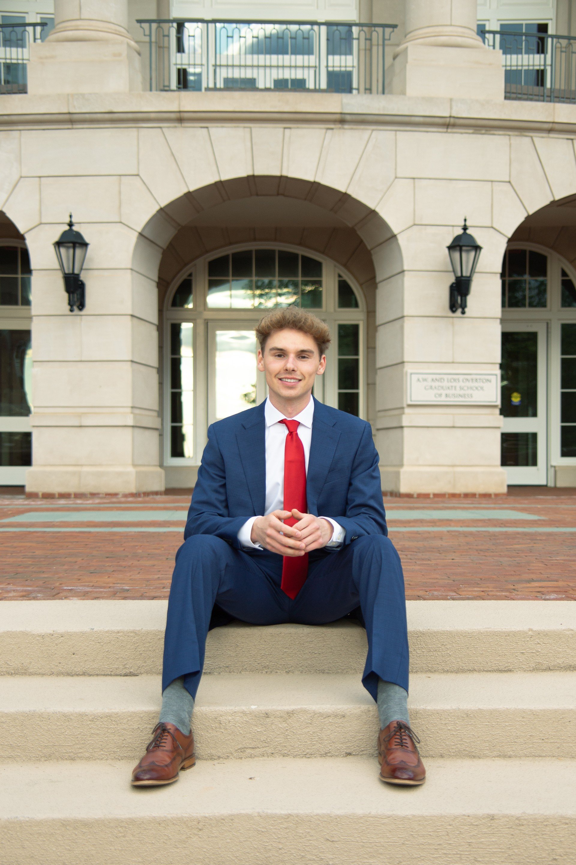 A young man in a blue suit with a red tie and brown shoes is sitting on stairs outside a building with large arched windows and stone walls, smiling at the camera.