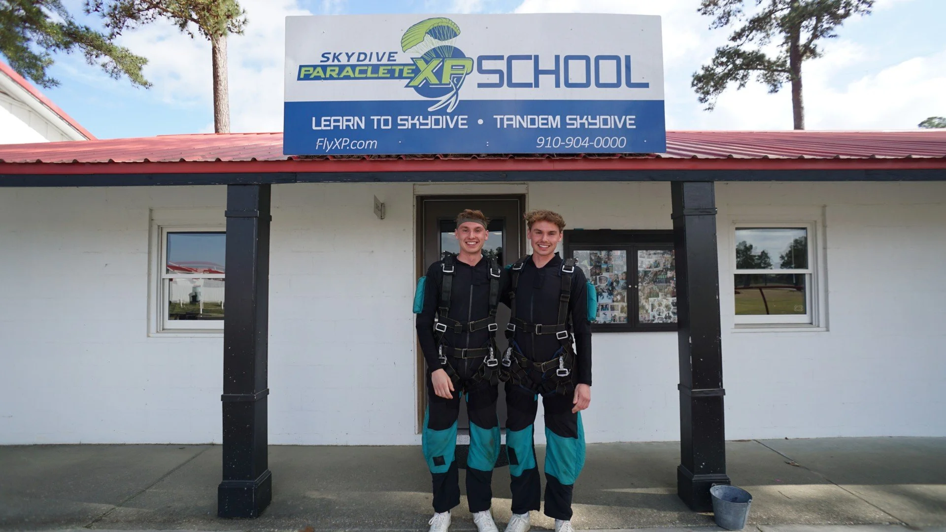 Two young men dressed in skydiving jumpsuits standing in front of a skydiving school building, smiling at the camera.