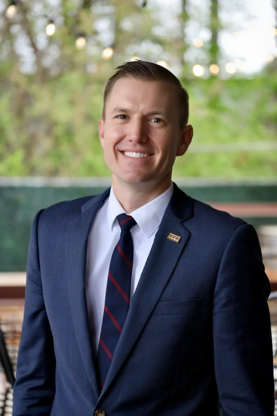 A young man with short brown hair, dressed in a navy blue suit, white shirt, and a striped tie, smiling in an outdoor setting with green trees and string lights in the background.