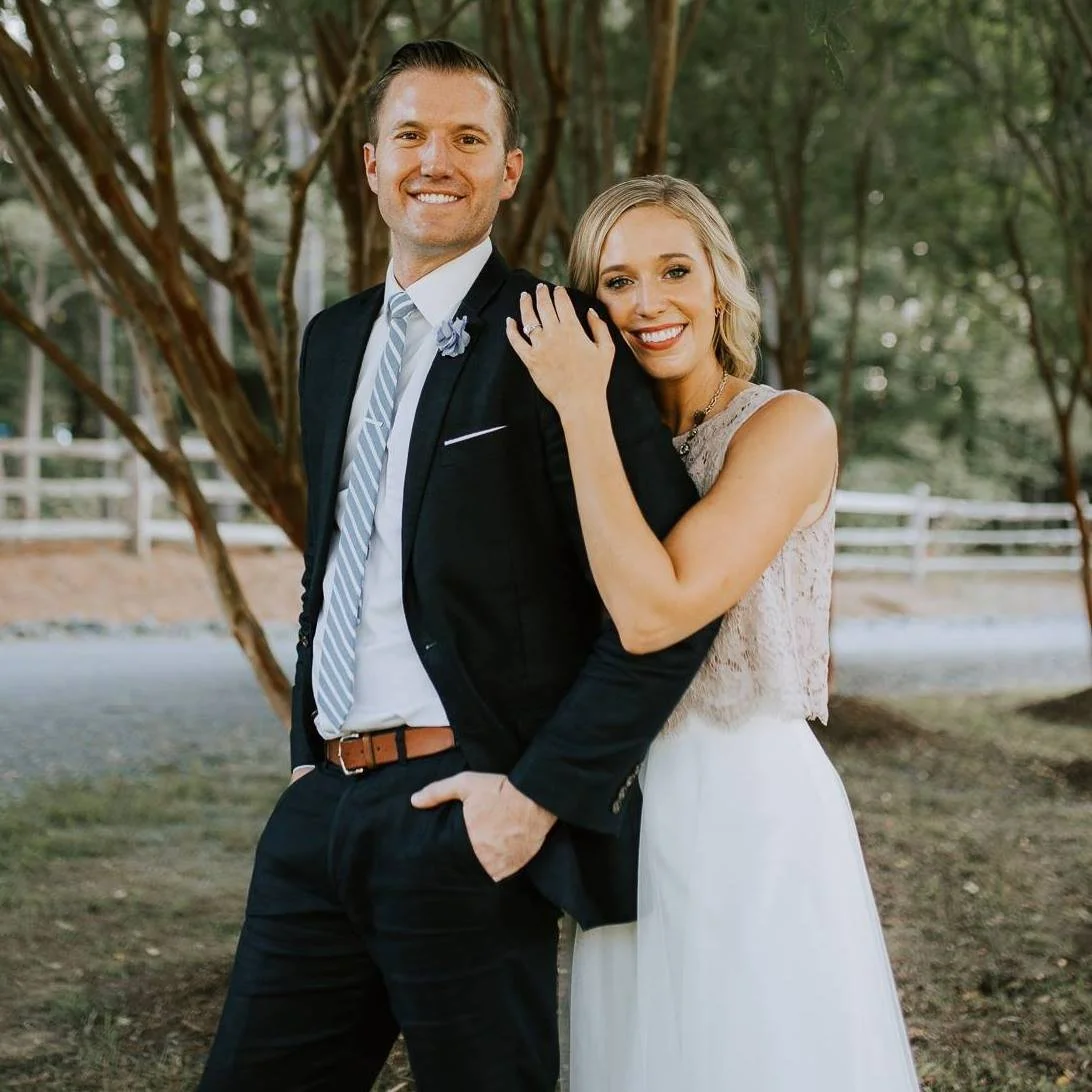 A smiling couple outdoors during daytime, with the woman hugging the man from the side. The man is wearing a dark suit with a light-colored tie, and the woman is wearing a sleeveless white dress with lace details. They are standing in front of trees and a white fence.