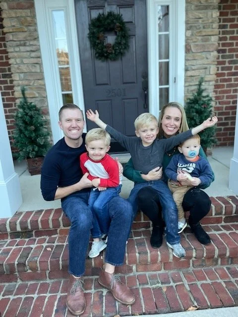 Family of four sitting on a brick porch in front of a door decorated with a holiday wreath, flanked by two small Christmas trees.