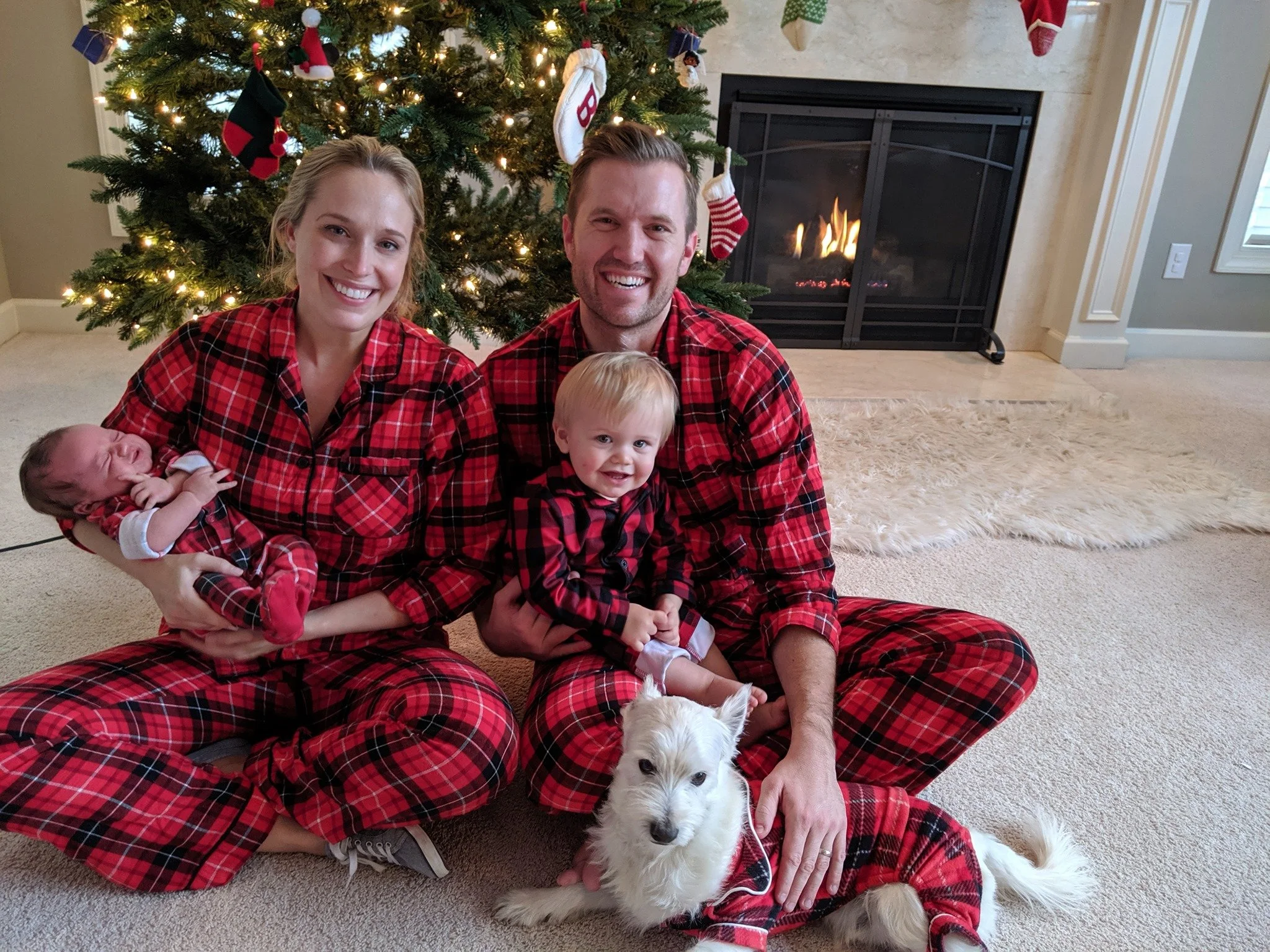 Family of four in matching red plaid pajamas sitting on a carpet in front of a decorated Christmas tree and fireplace. The mother is holding a newborn, the father is holding their toddler, and a white dog is laying on the floor in front of them.