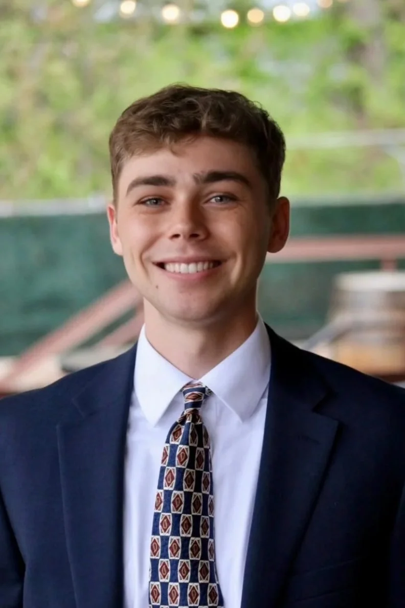 A young man smiling, dressed in a dark suit, white shirt, and patterned tie, with a blurred outdoor background.