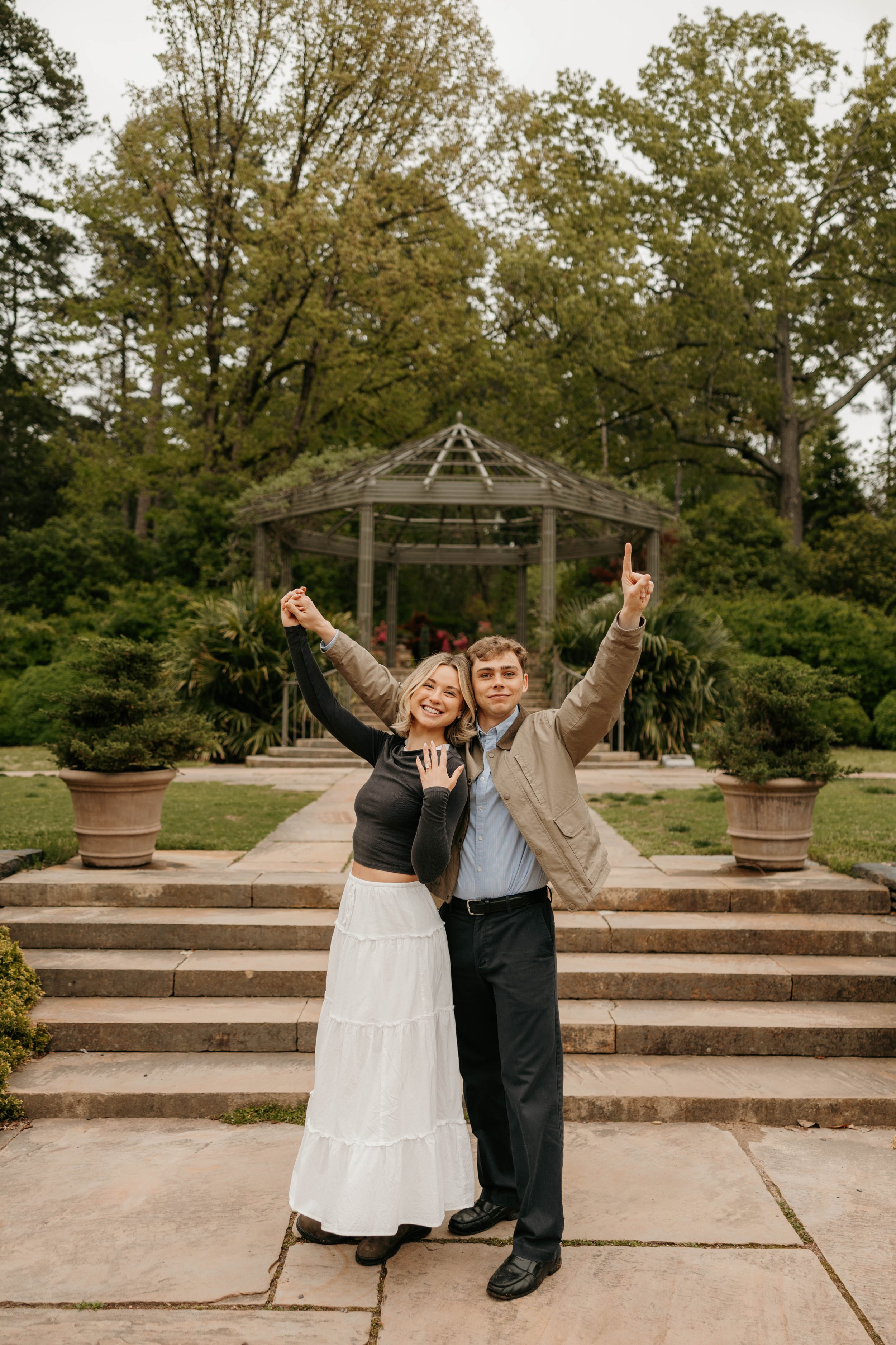 A young woman and young man are celebrating outdoors on steps in front of a gazebo. They are smiling, with the woman wearing a black long-sleeve top and a white skirt, and the man wearing a beige jacket and dark pants. The woman has her hand on her chest, and the man is raising one finger in the air.