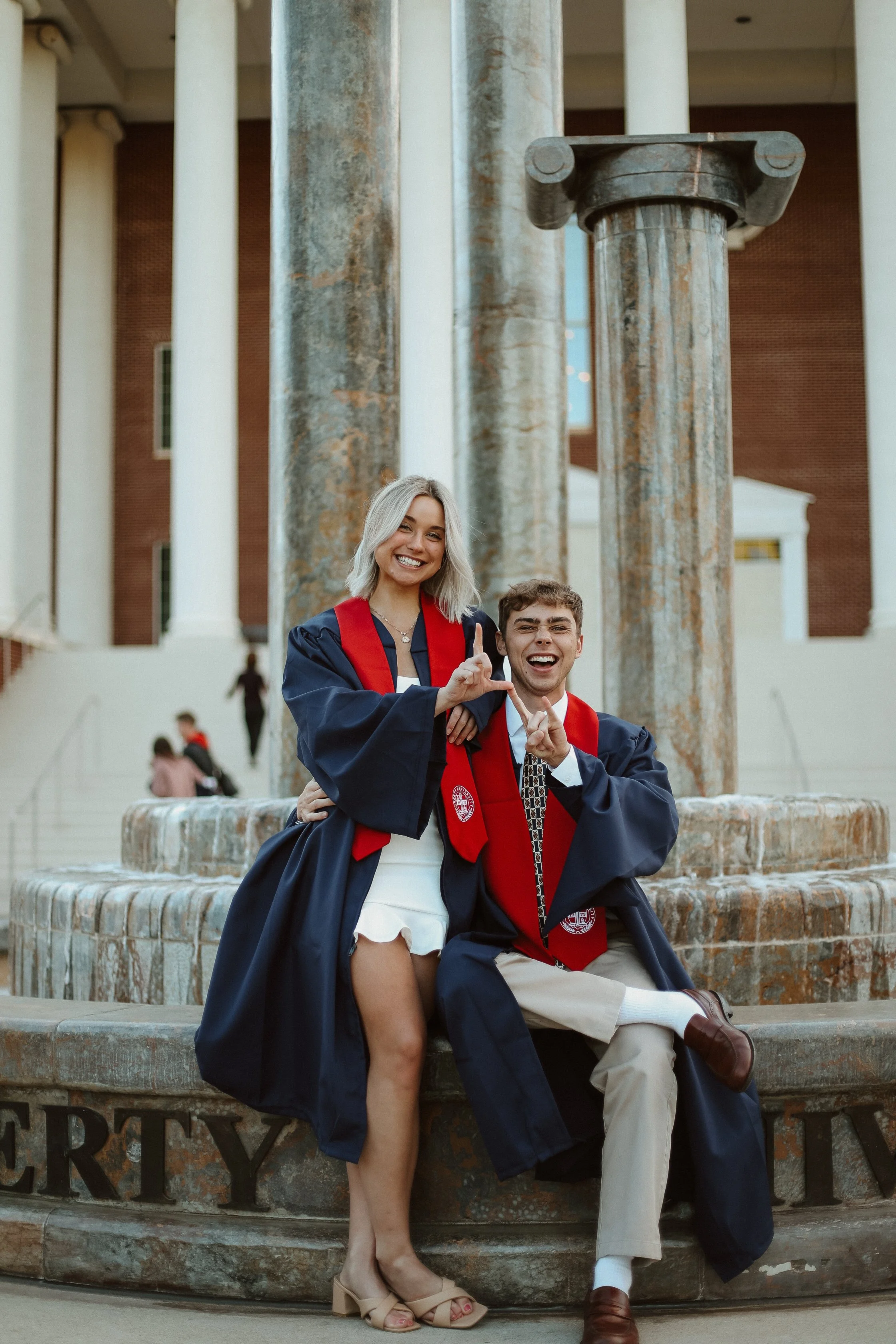 Two graduates in caps and gowns sitting on a stone fountain, celebrating and making hand signs in front of a large building with columns.