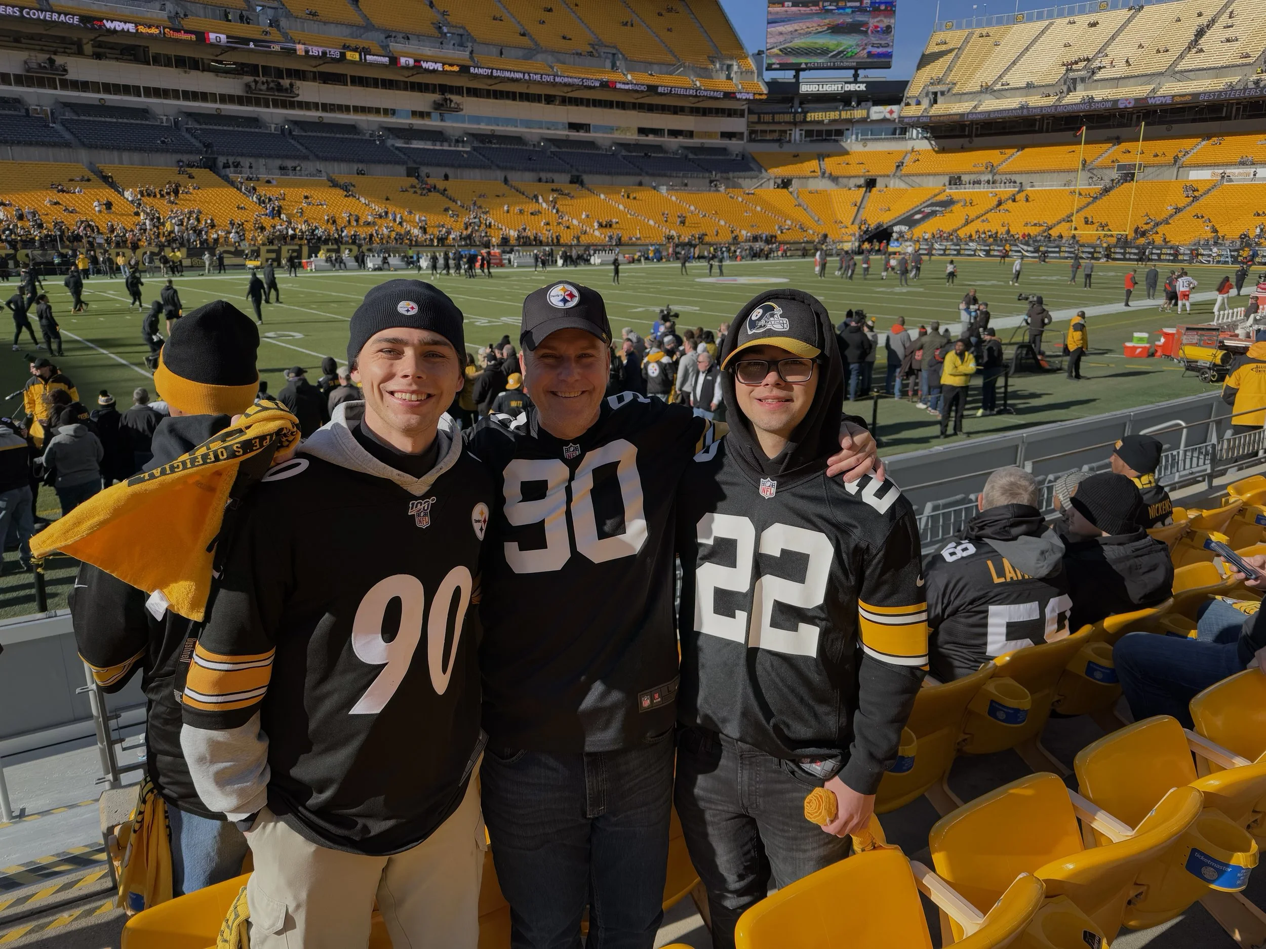 Three people standing in front of a football field at Heinz Field in Pittsburgh, wearing black and yellow Pittsburgh Steelers jerseys, smiling, with fans and players in the background.