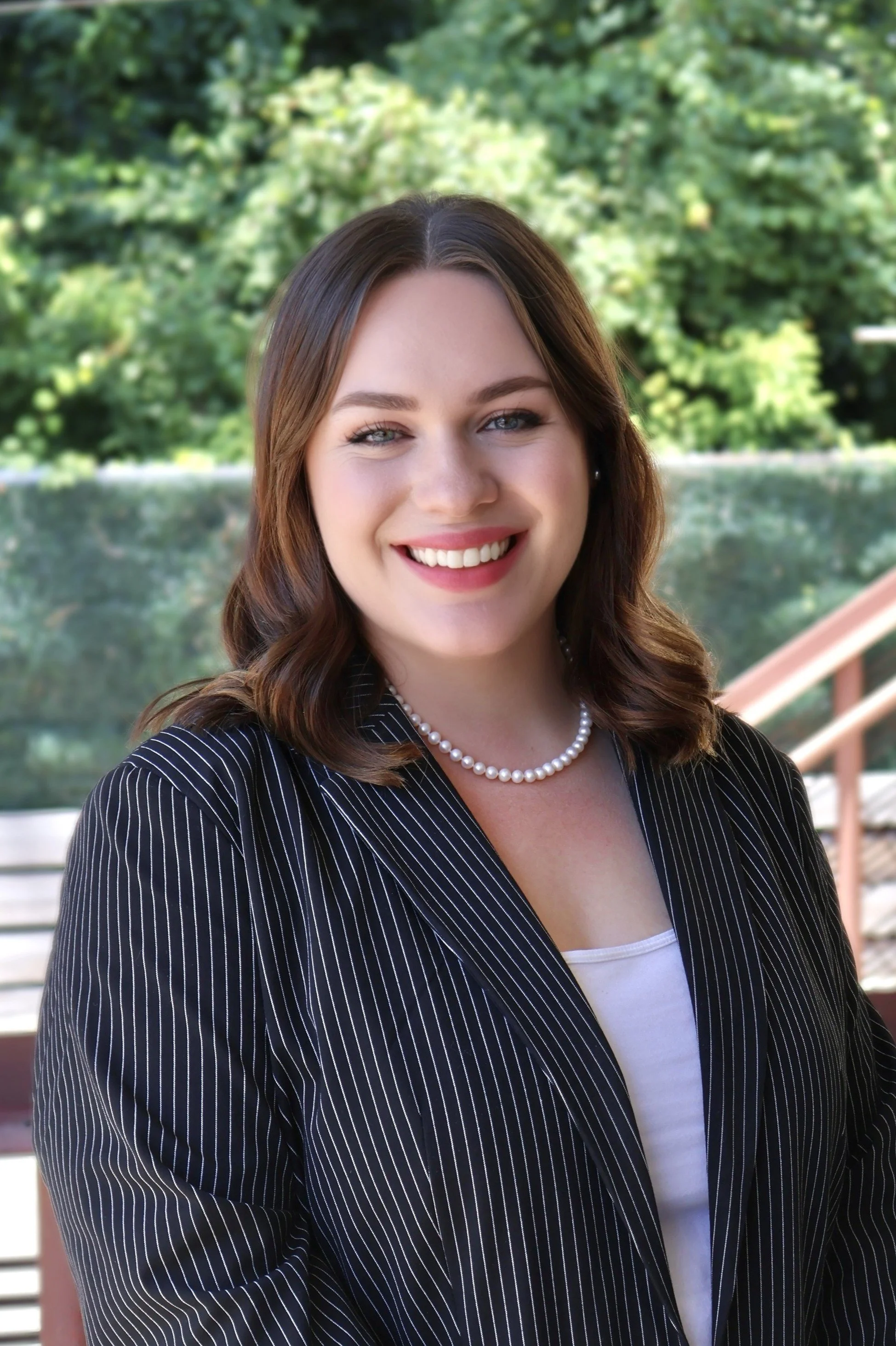 A young woman smiling outdoors with a background of trees and a wooden railing, wearing a black pinstripe blazer, a white top, a pearl necklace, and earrings.