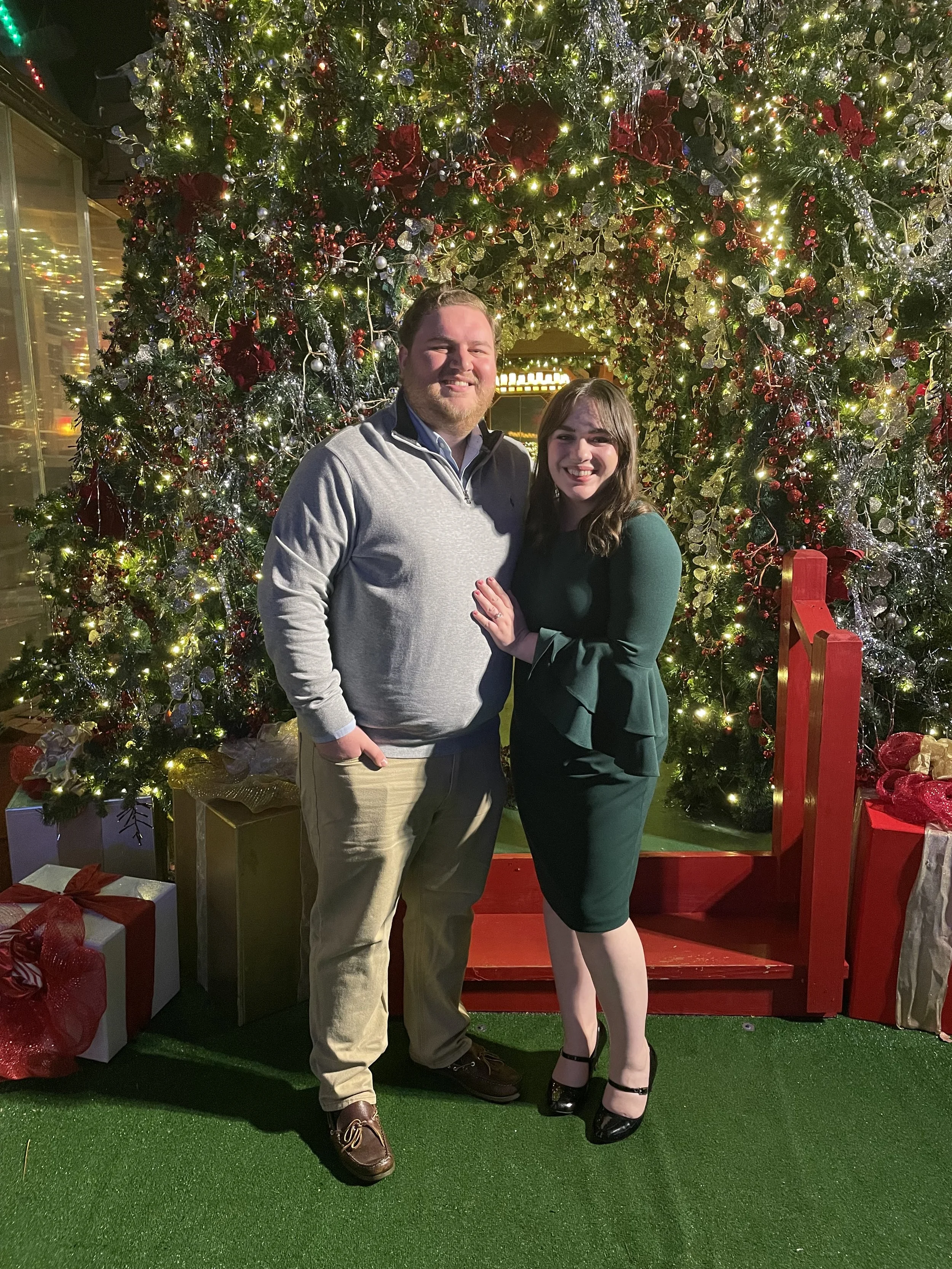 A man and woman pose together outdoors at night in front of a large decorated Christmas tree with lights and ornaments, surrounded by wrapped presents, under a festive archway.