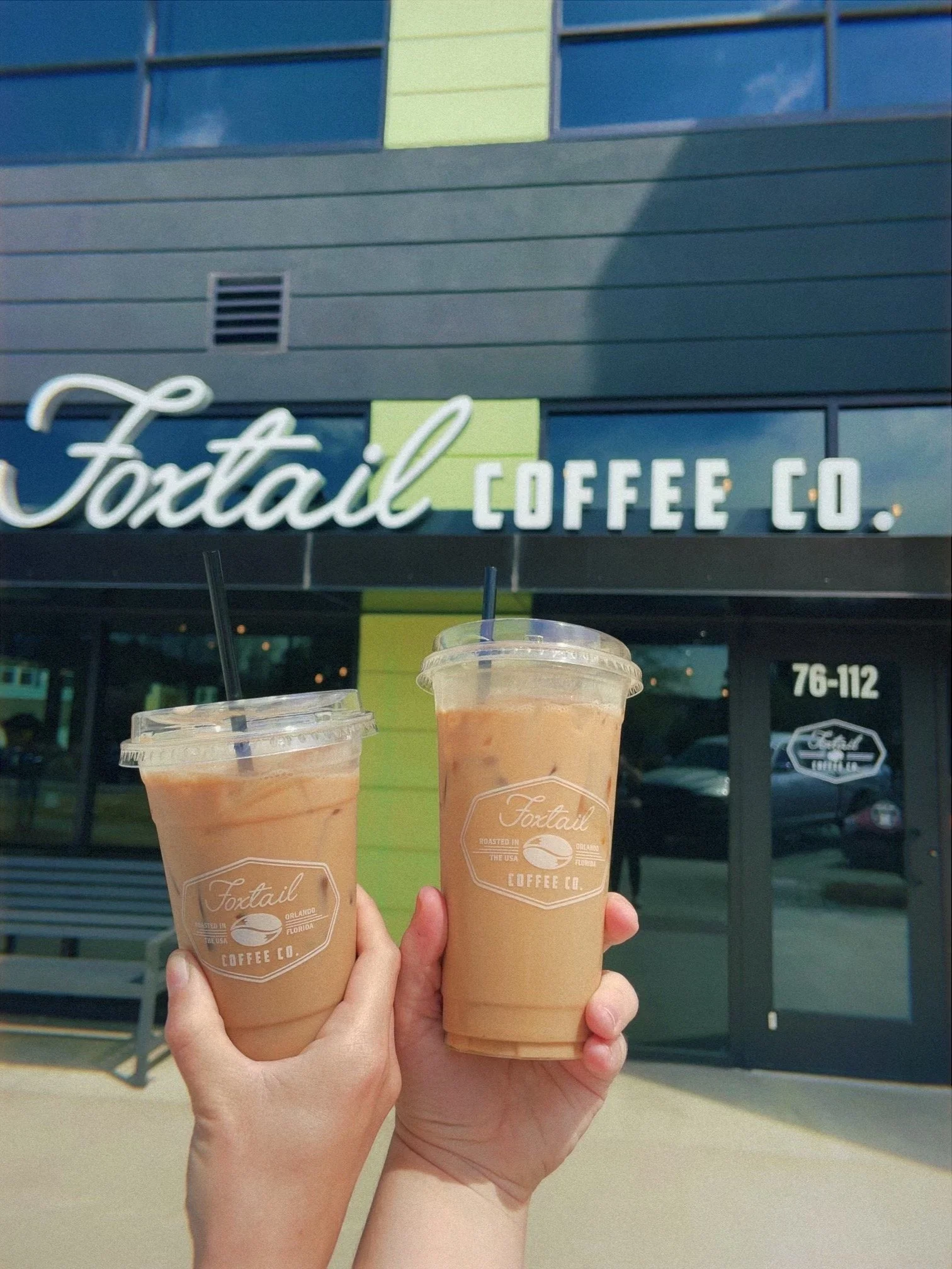 Two hands holding iced coffee drinks with black straws in front of the Foxtail Coffee Co. storefront.