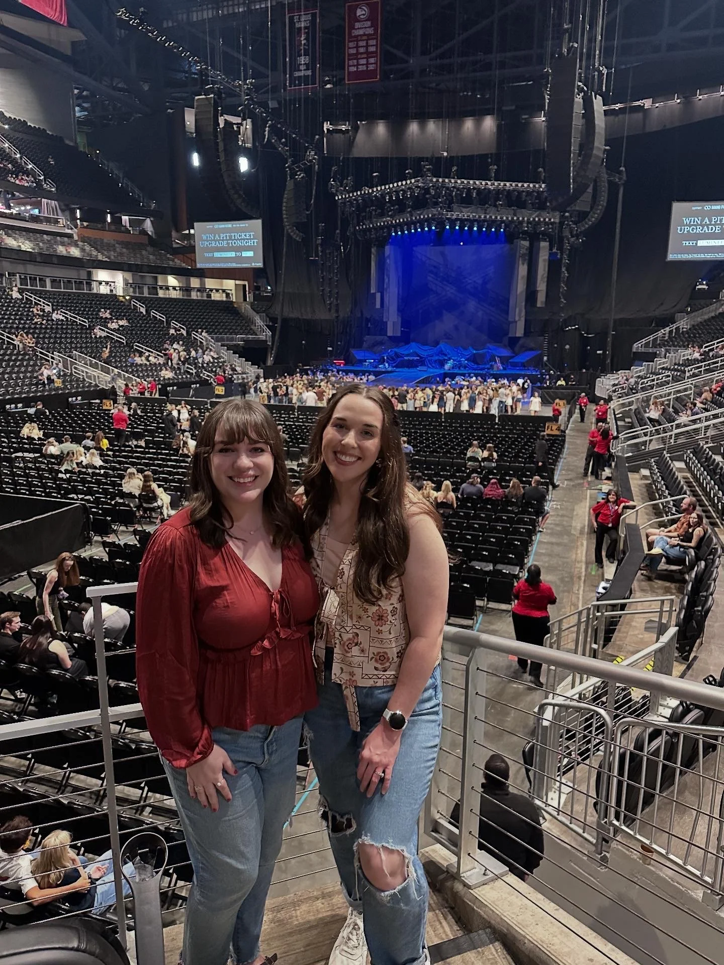 Two women smiling at a concert arena with empty seats and a stage in the background.