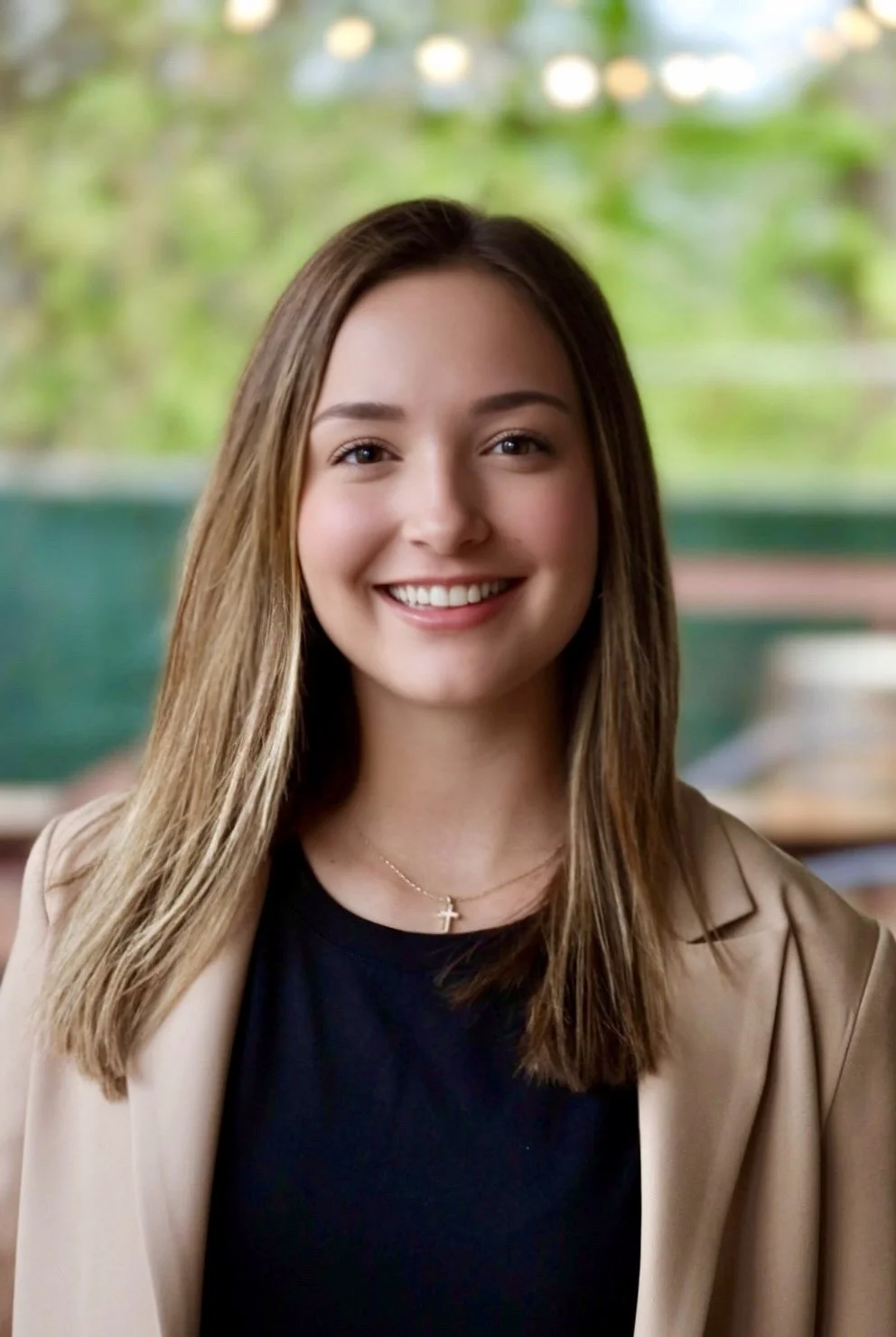 A young woman with straight, shoulder-length brown hair and a warm smile, wearing a black top, beige blazer, and a gold necklace with a cross pendant, standing in front of a blurred green outdoor setting.