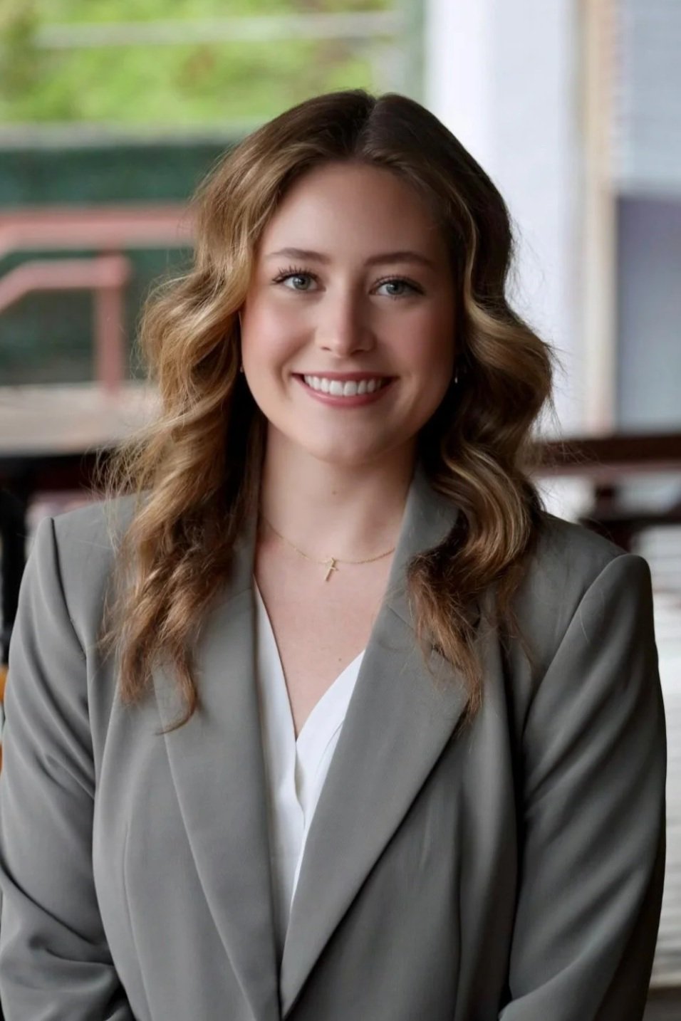 A woman with wavy, light brown hair and blue eyes smiling, wearing a gray blazer and a small gold cross necklace, sitting indoors with a blurred background of a classroom or office.