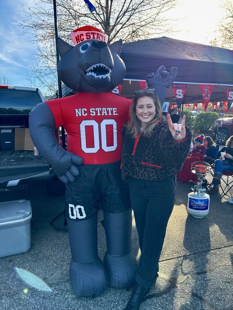 A woman smiling and making a rock hand sign standing next to a giant inflatable mascot of a black wolf wearing NC State gear, including a red hat and red jersey with the number 00, at a tailgate or outdoor event with other people and flags in the background.