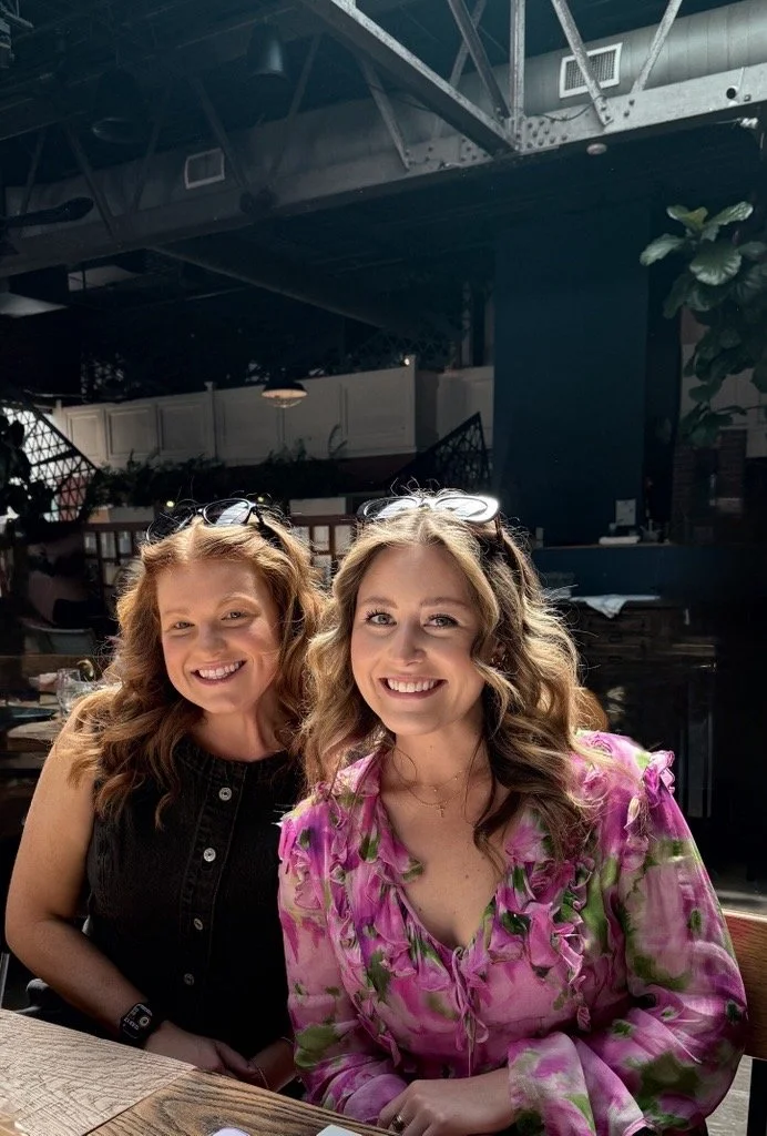 Two women with wavy hair and sunglasses on their heads smiling at a restaurant table.