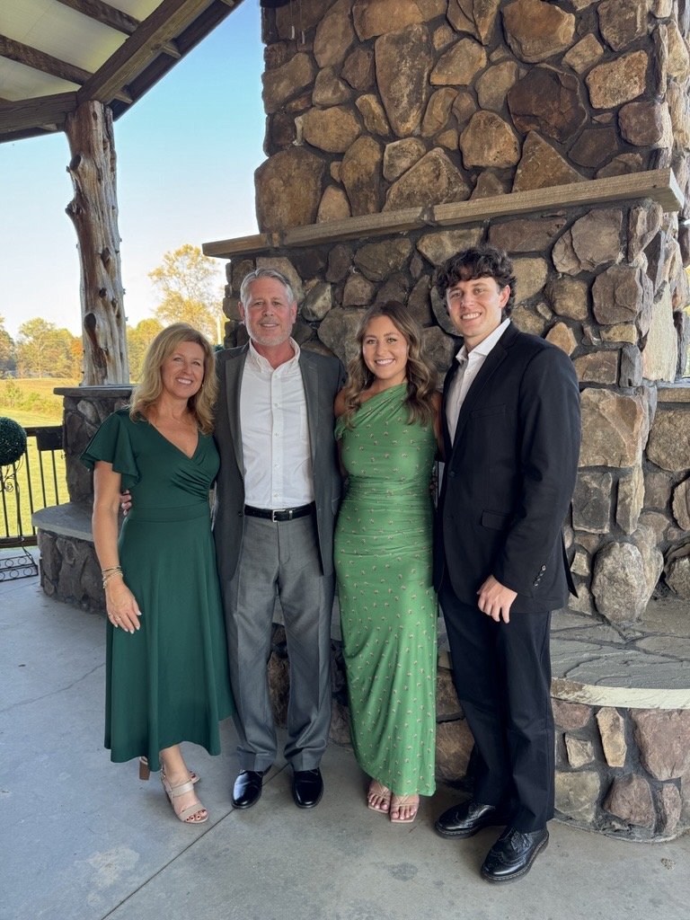Four people dressed in formal attire standing together in front of a large stone fireplace on a covered porch, with a scenic outdoor background.