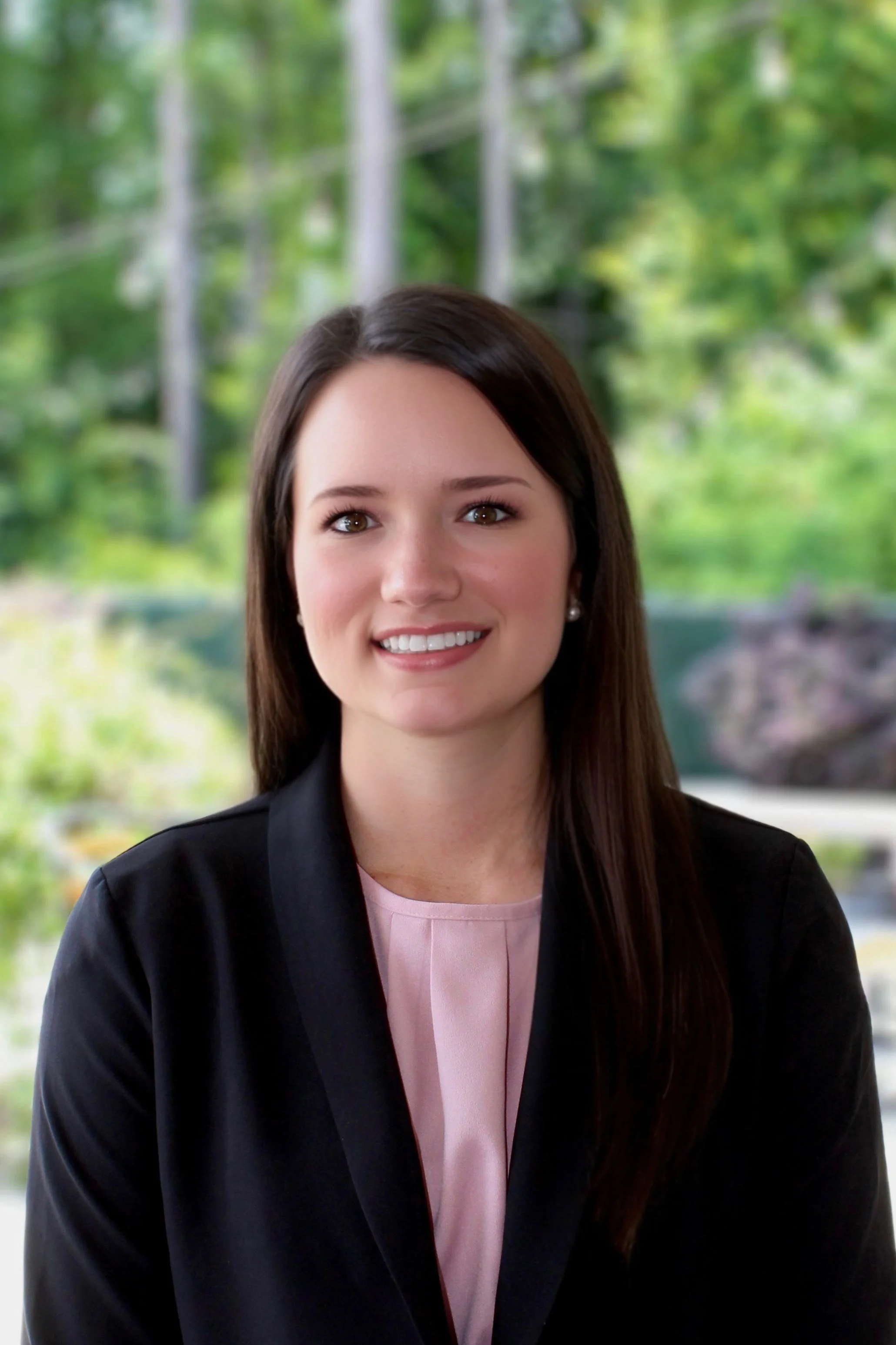A woman with long brown hair smiling, wearing a black blazer and pink blouse, sitting outdoors with a blurred green, wooded background.