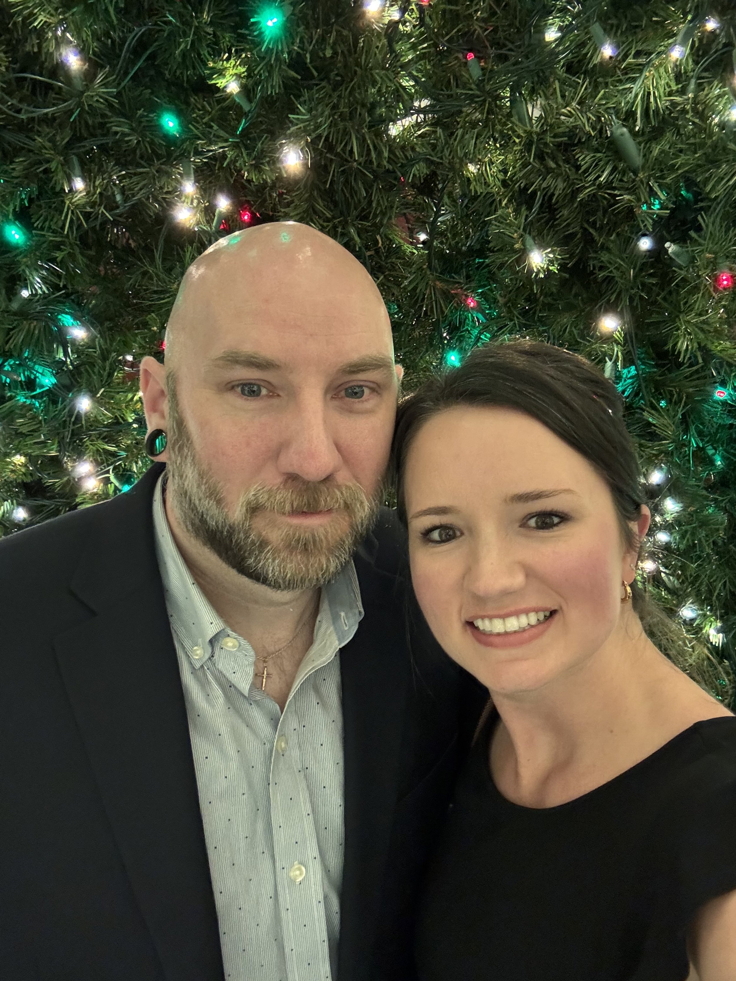 A man and a woman taking a selfie in front of a decorated Christmas tree with colorful lights.
