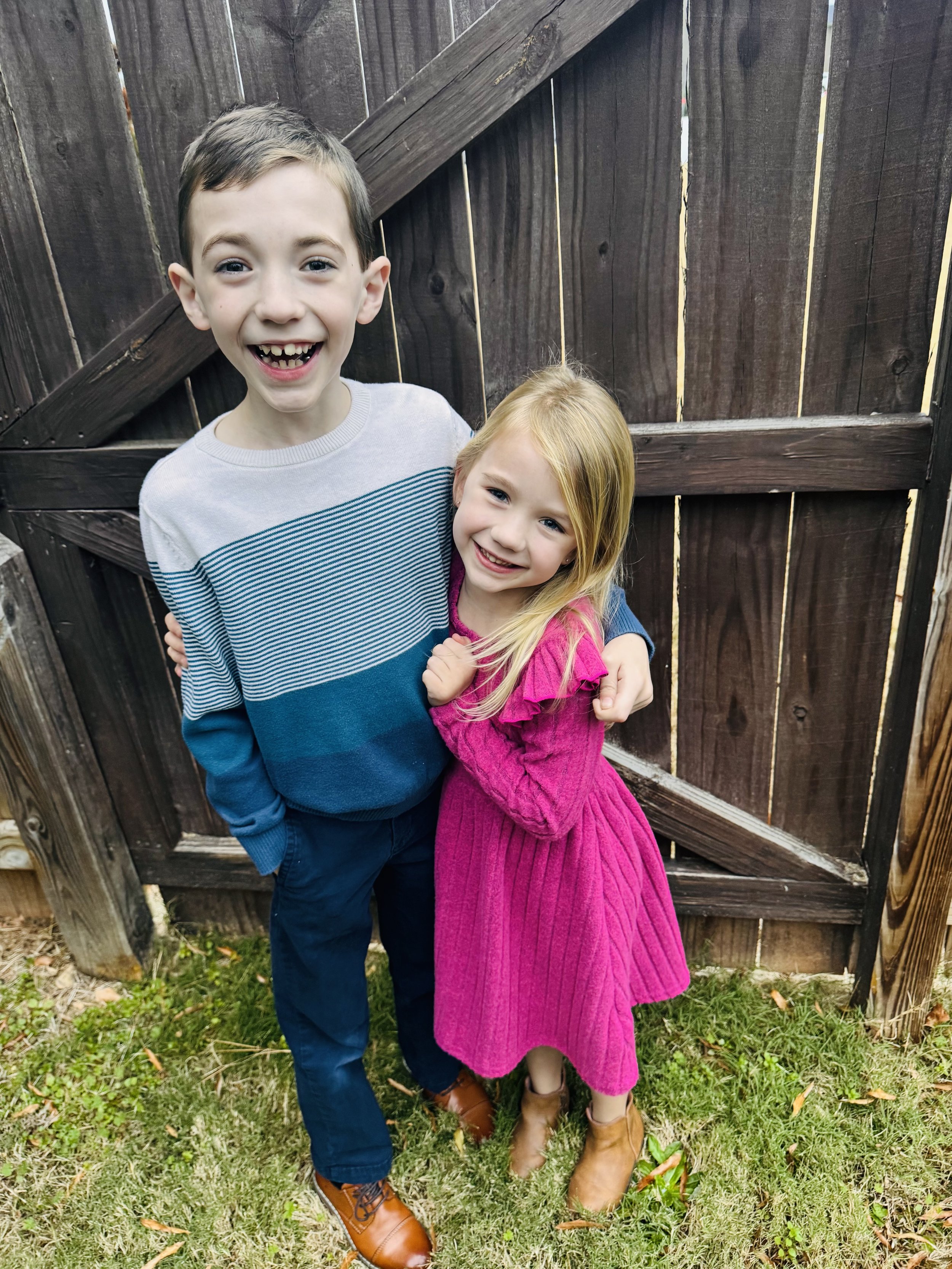 Two children, a boy and a girl, smiling and hugging each other outdoors in front of a wooden fence.