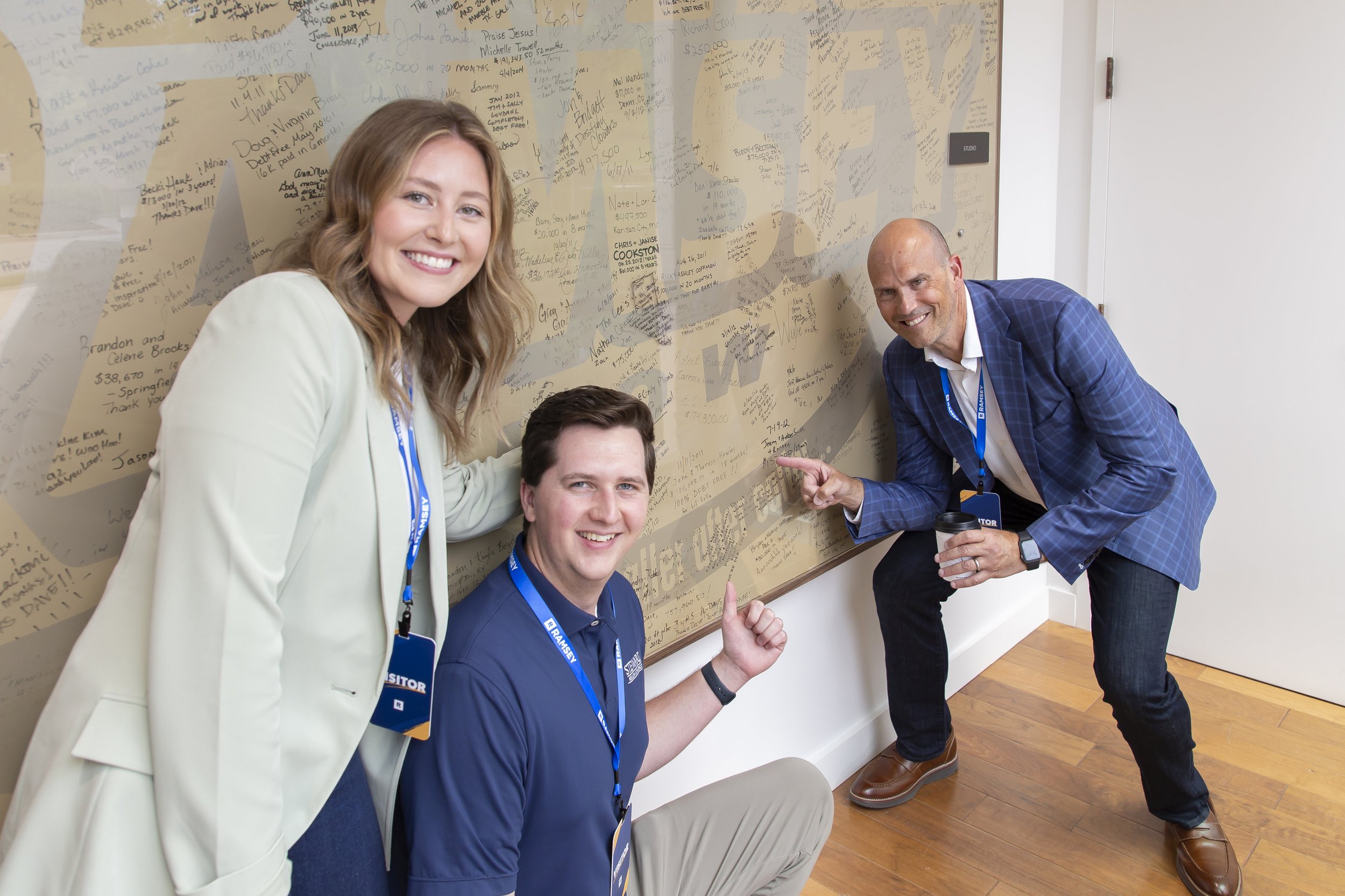 Three smiling individuals, two men and a woman, pointing at and standing near a wall covered with handwritten notes and messages, in a professional setting with light-colored walls and wooden floors.