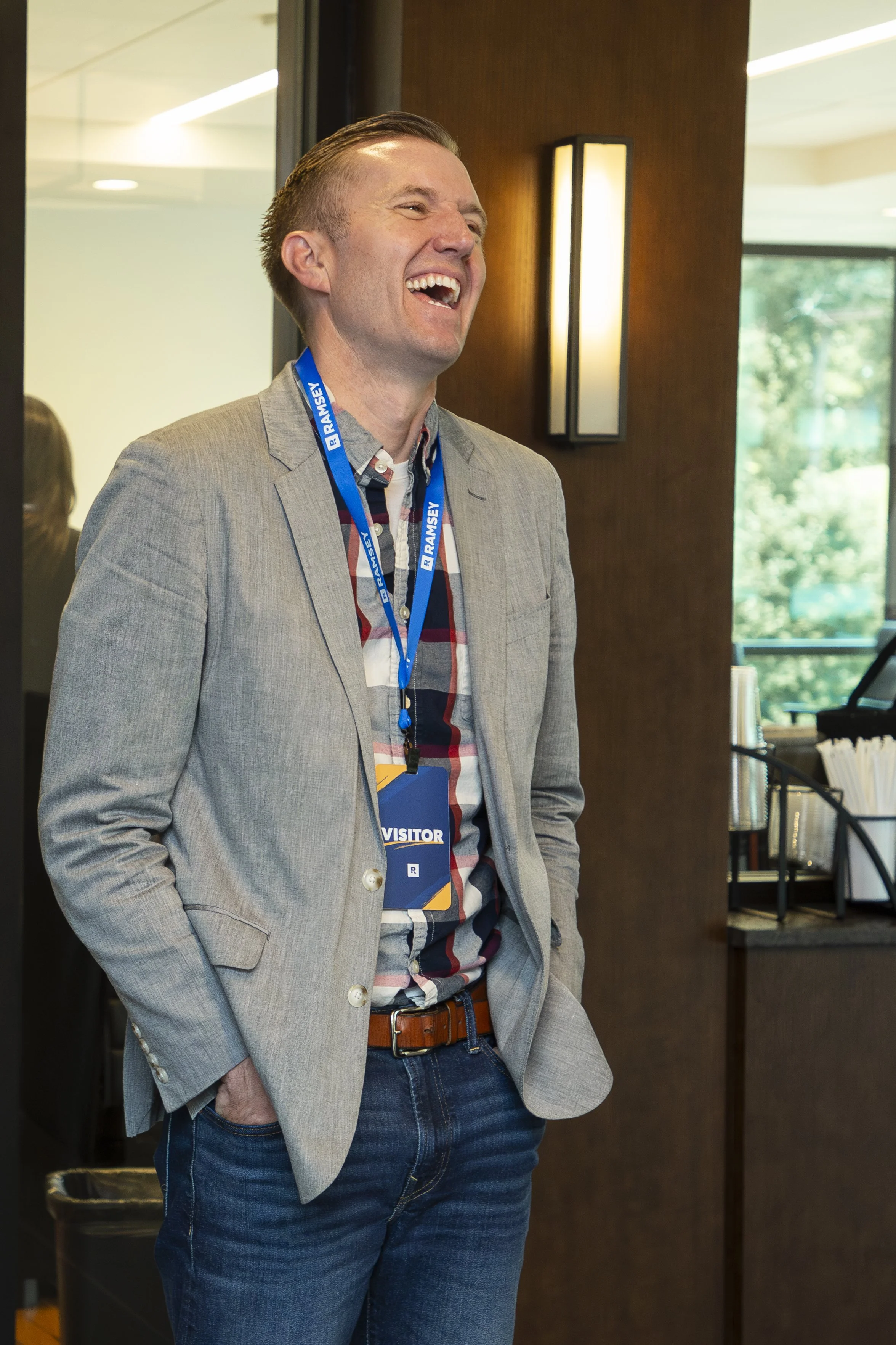 A man laughing and smiling while standing indoors at a conference or event, wearing a gray blazer, a lanyard with a visitor badge, and casual jeans.