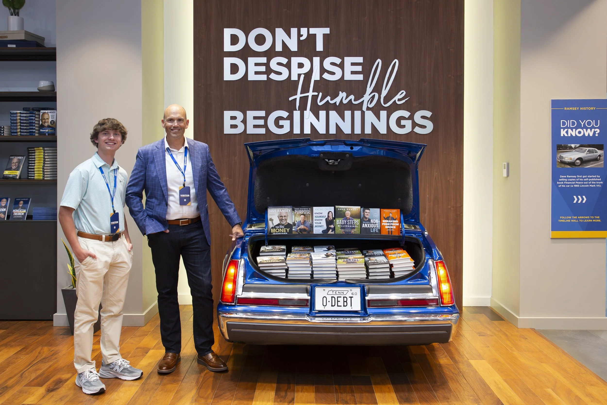 Two men standing inside office with a vintage car trunk filled with books and a large sign behind them that reads 'Don't Despise Humble Beginnings'.