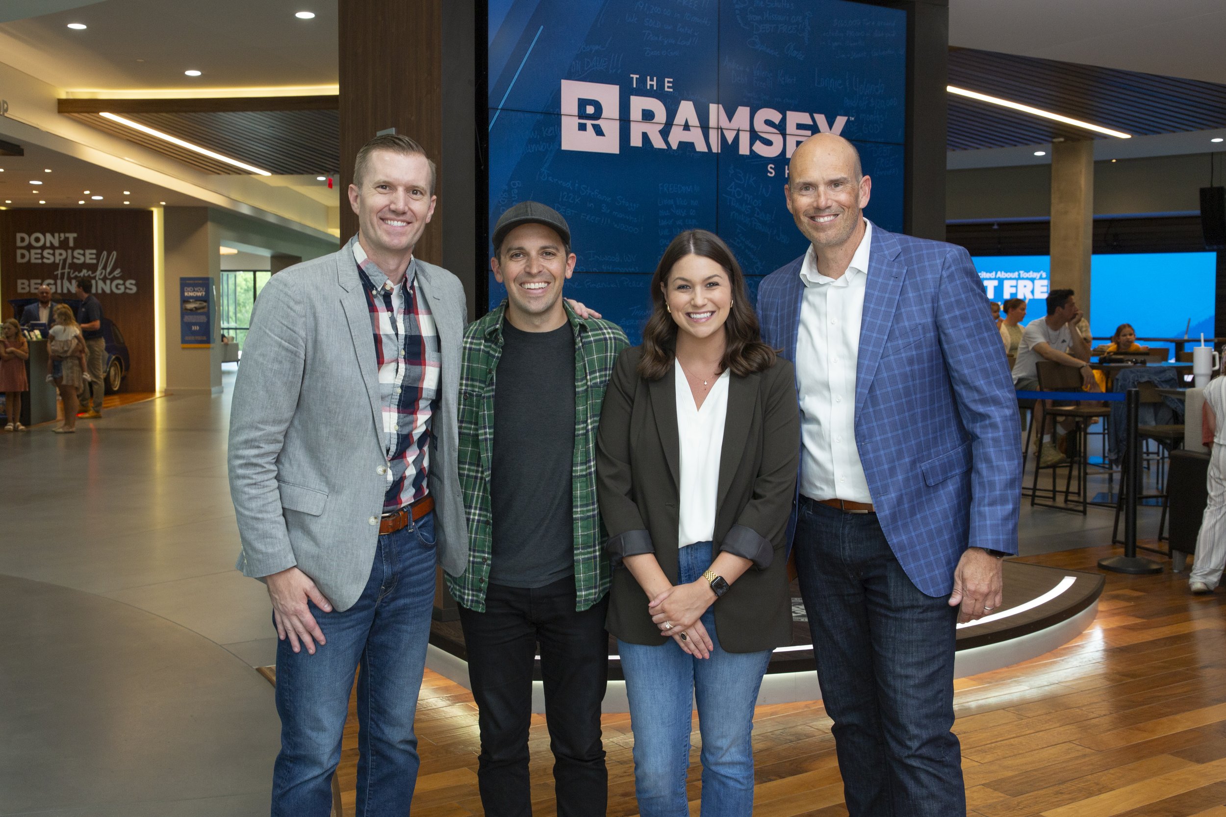 Group of four people standing and smiling in front of a digital screen displaying 'The Ramsey Show' in an indoor setting, likely a conference or event center.