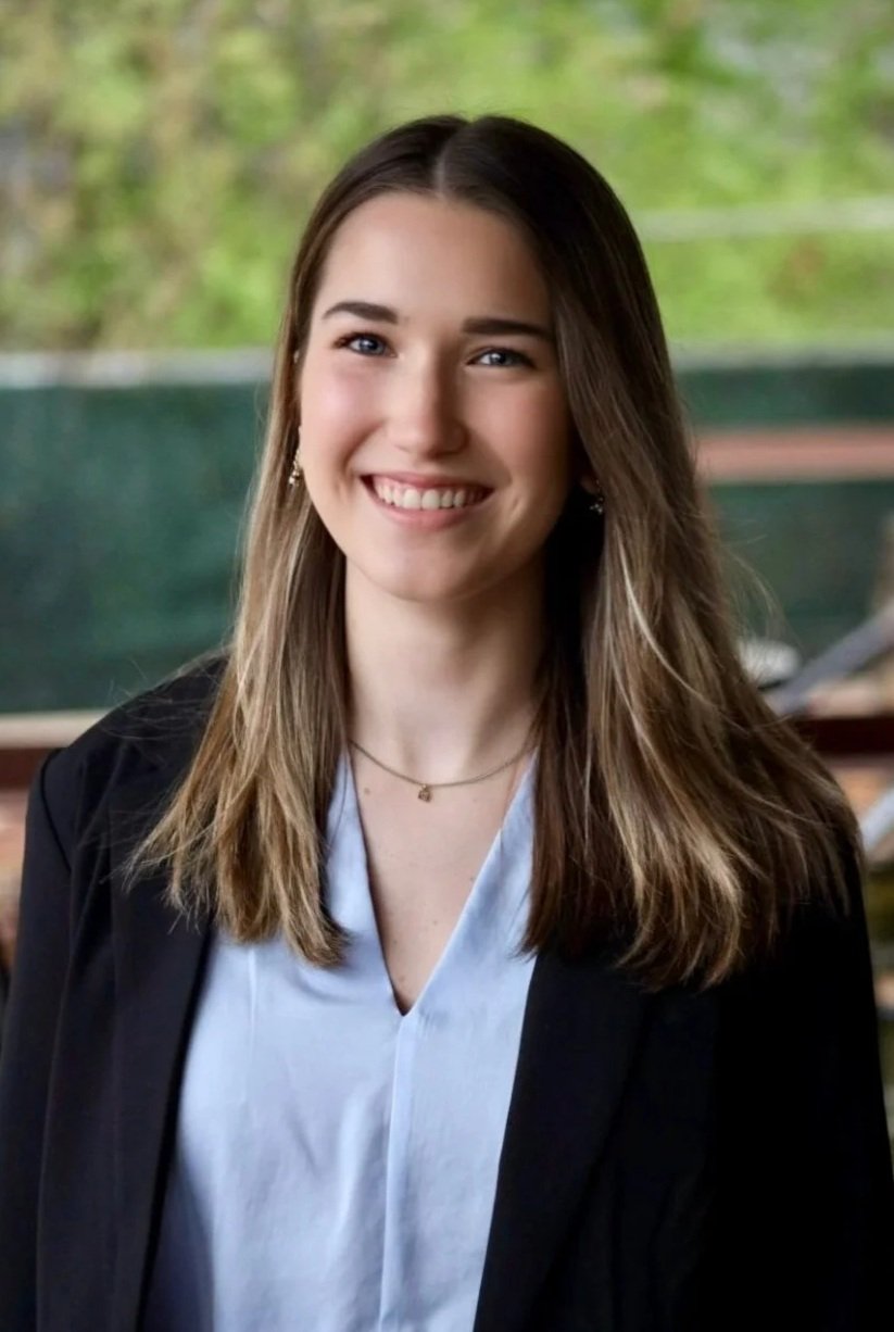 A young woman with long brown hair and blue eyes, smiling, wearing a black blazer over a white blouse, standing outdoors with greenery in the background.