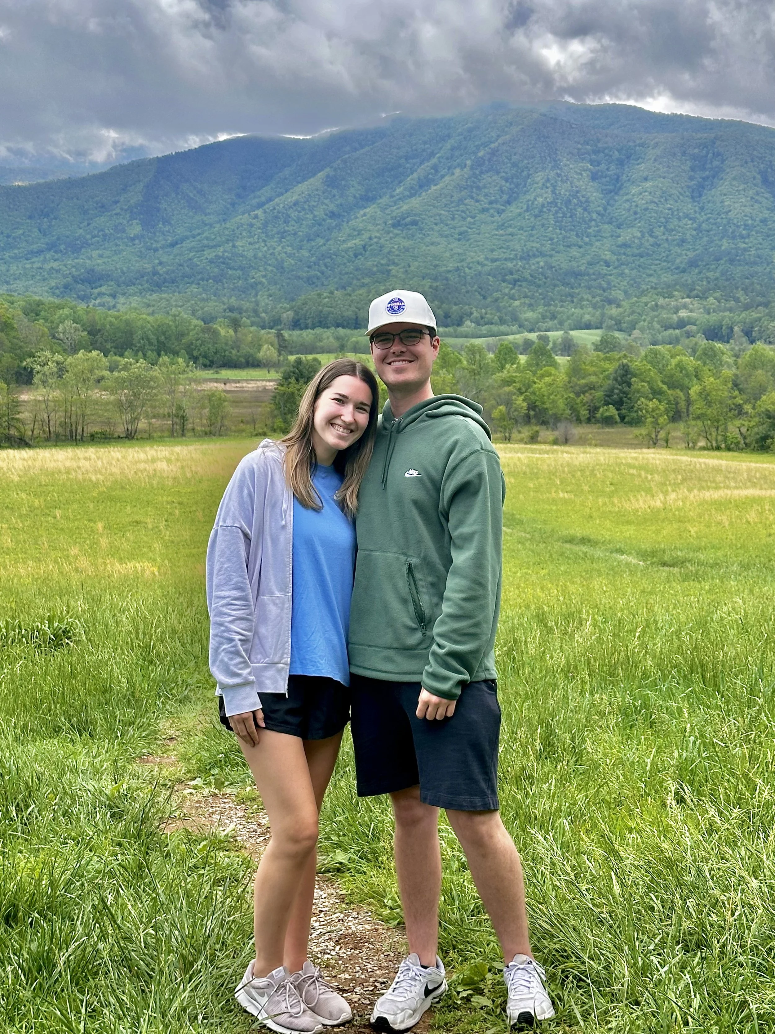 A young couple standing on a dirt path in a green field with a mountain and cloudy sky in the background. They are smiling and embracing.
