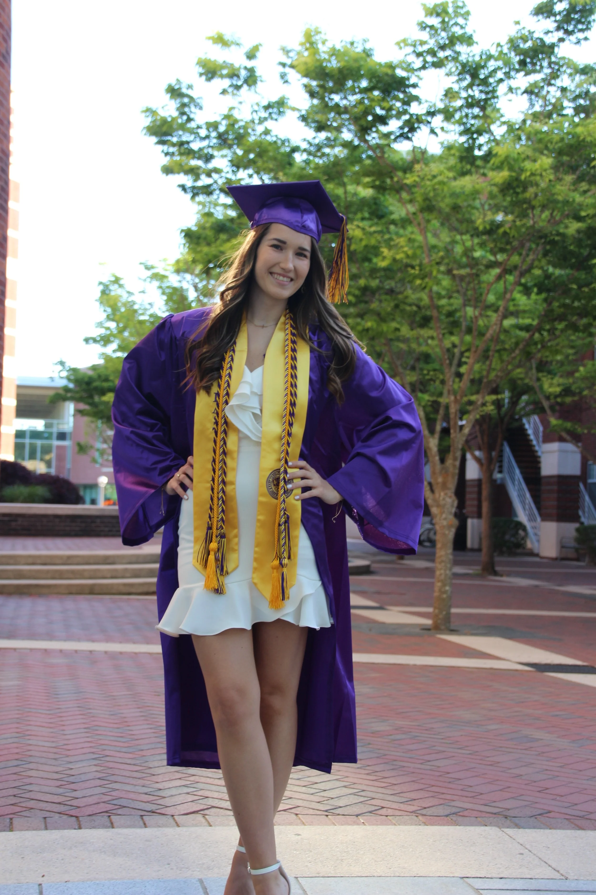 Young woman in purple graduation gown and cap, smiling outside on a college campus, wearing honor cords and medals.