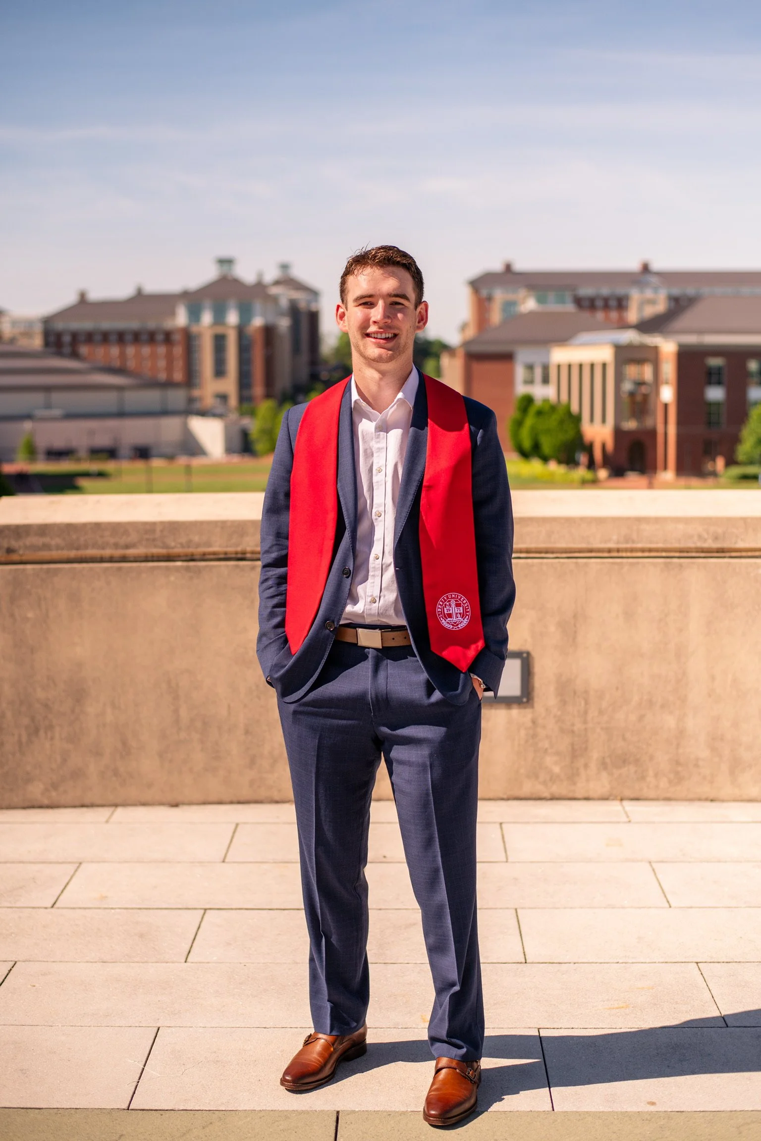 Young man in a suit with a red graduation stole standing outdoors with university buildings in the background.