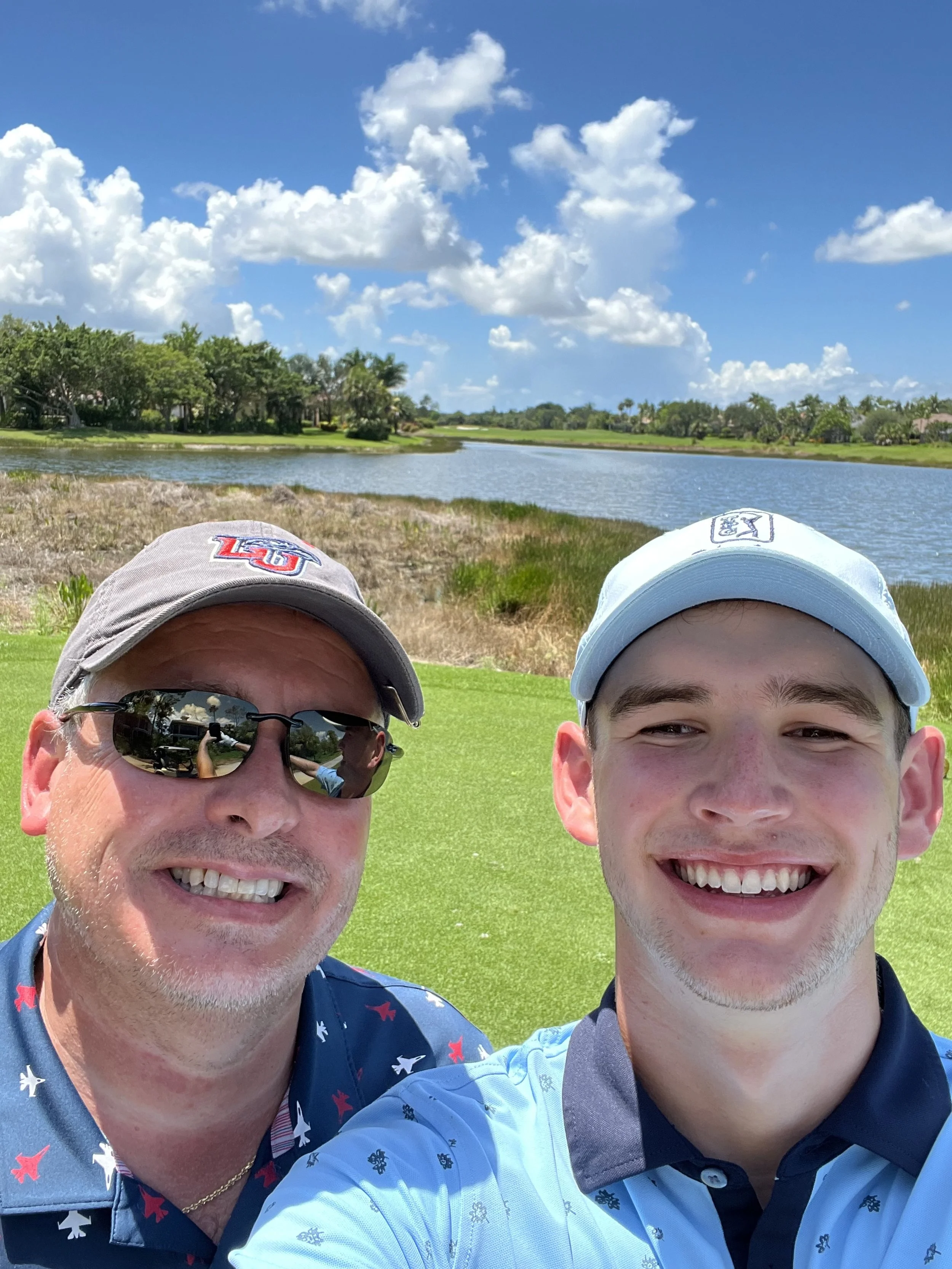 Two men stand on a golf course near a pond with green grass, trees, and a partly cloudy sky in the background, smiling for a selfie.
