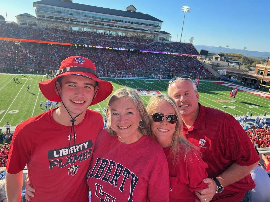 Photo of a family at a football game, wearing Liberty Flames team apparel, in a stadium filled with fans, with a football field and stadium structures in the background.