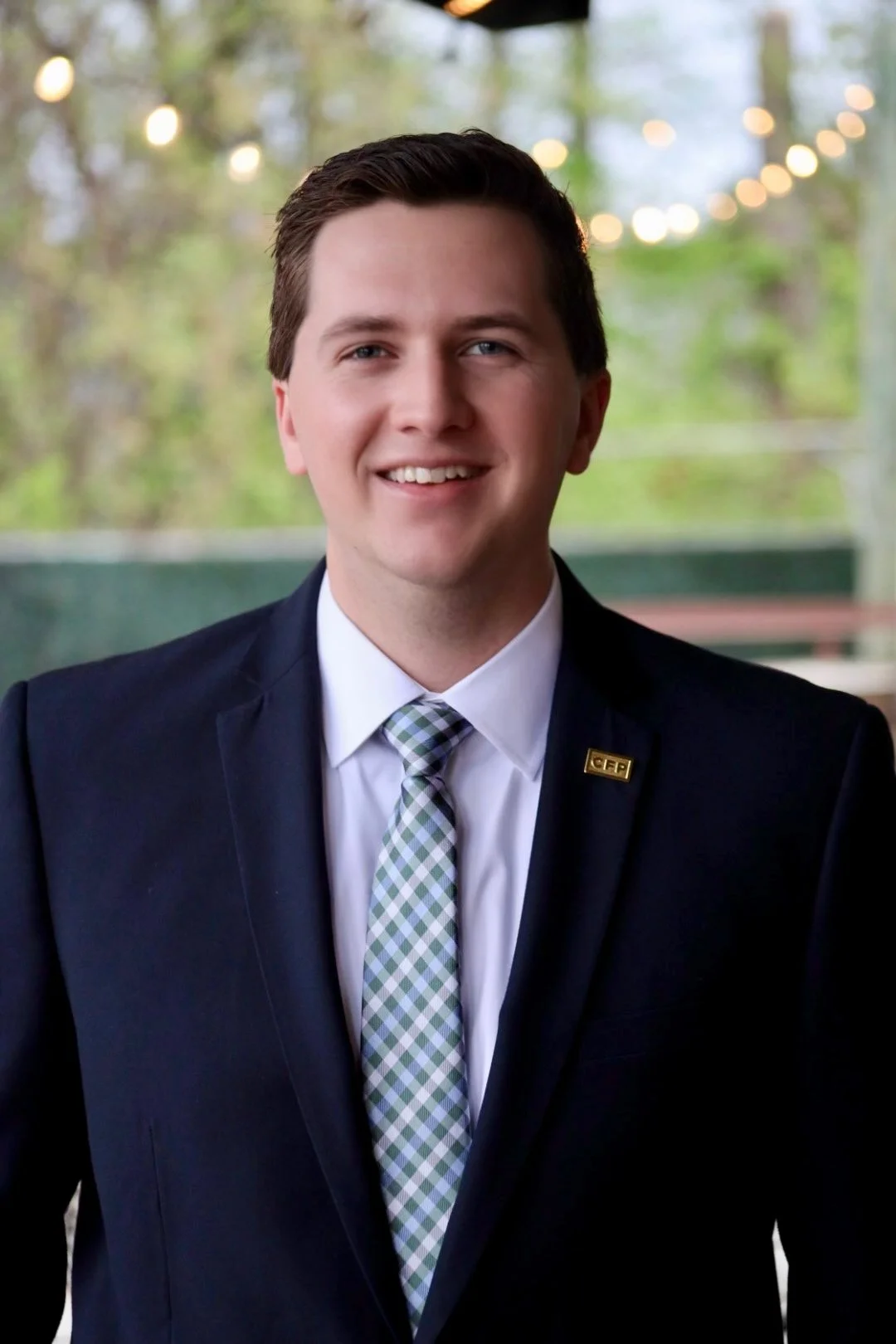 A young man with short brown hair, wearing a dark suit, white shirt, checkered tie, and a pin on his lapel, standing outdoors with blurred string lights and trees in the background.